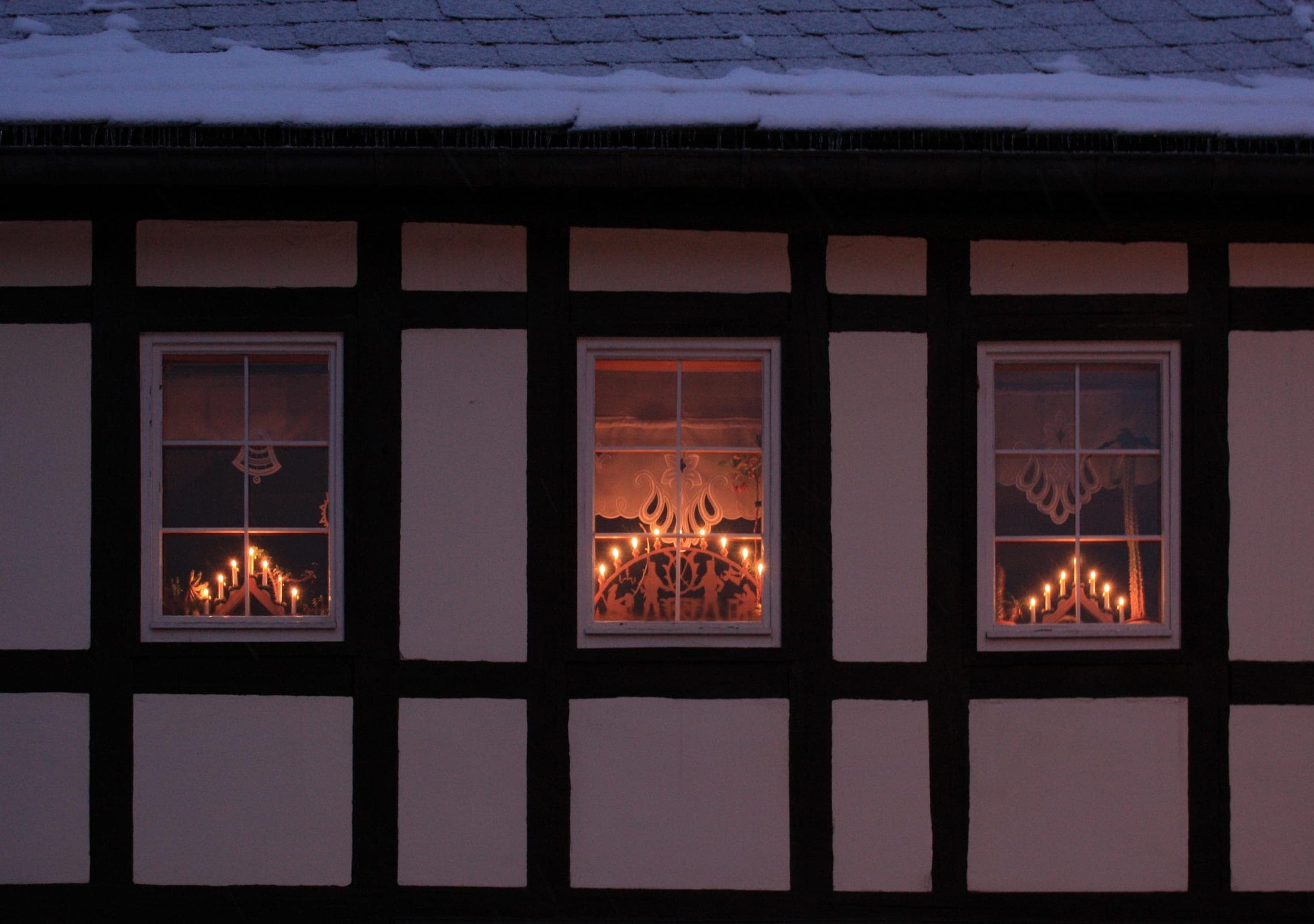 Candle arches glow in three windows of a half-timbered house in Rittersgrün in the Ore Mountains at dusk.