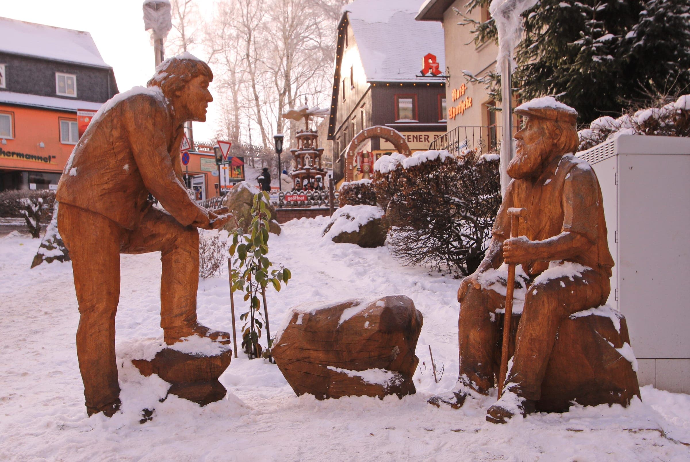 Two carved wooden figures sit in the snow on Seiffen's main street with a Christmas pyramid behind them.