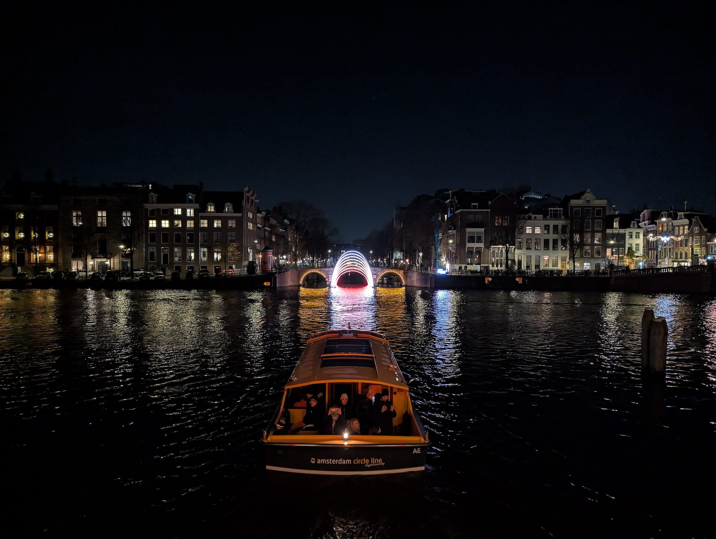 A canal boat crosses the Amstel towards a lit arch at the mouth of the Herengracht during Amsterdam Light Festival.