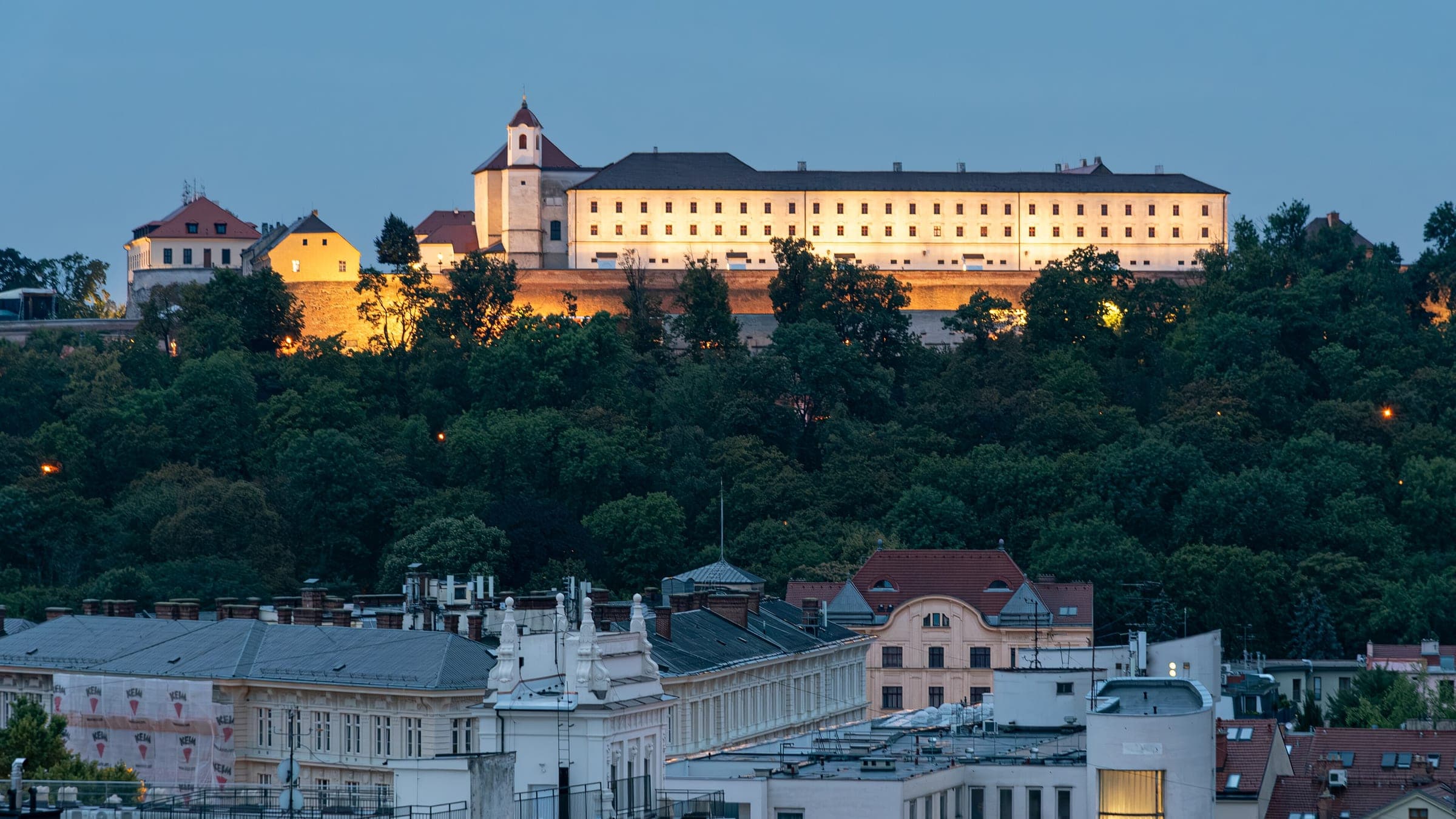 Floodlights come on along Špilberk Castle at blue hour on the wooded hill above Brno.