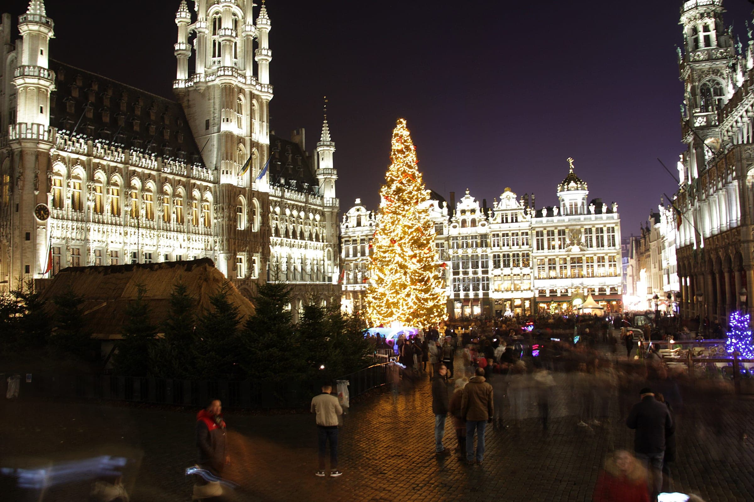 Visitors stand on the Grand-Place in Brussels at night under a tall Christmas tree strung with warm lights.