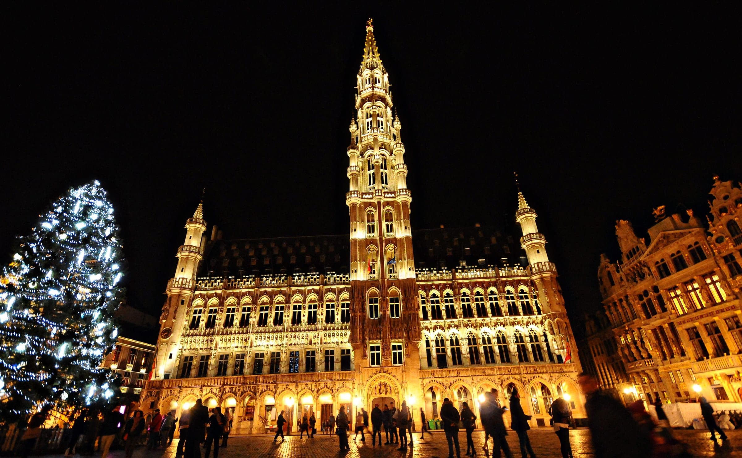 Crowds gather on the Grand-Place in Brussels at night beside a lit tree as the town hall glows gold.