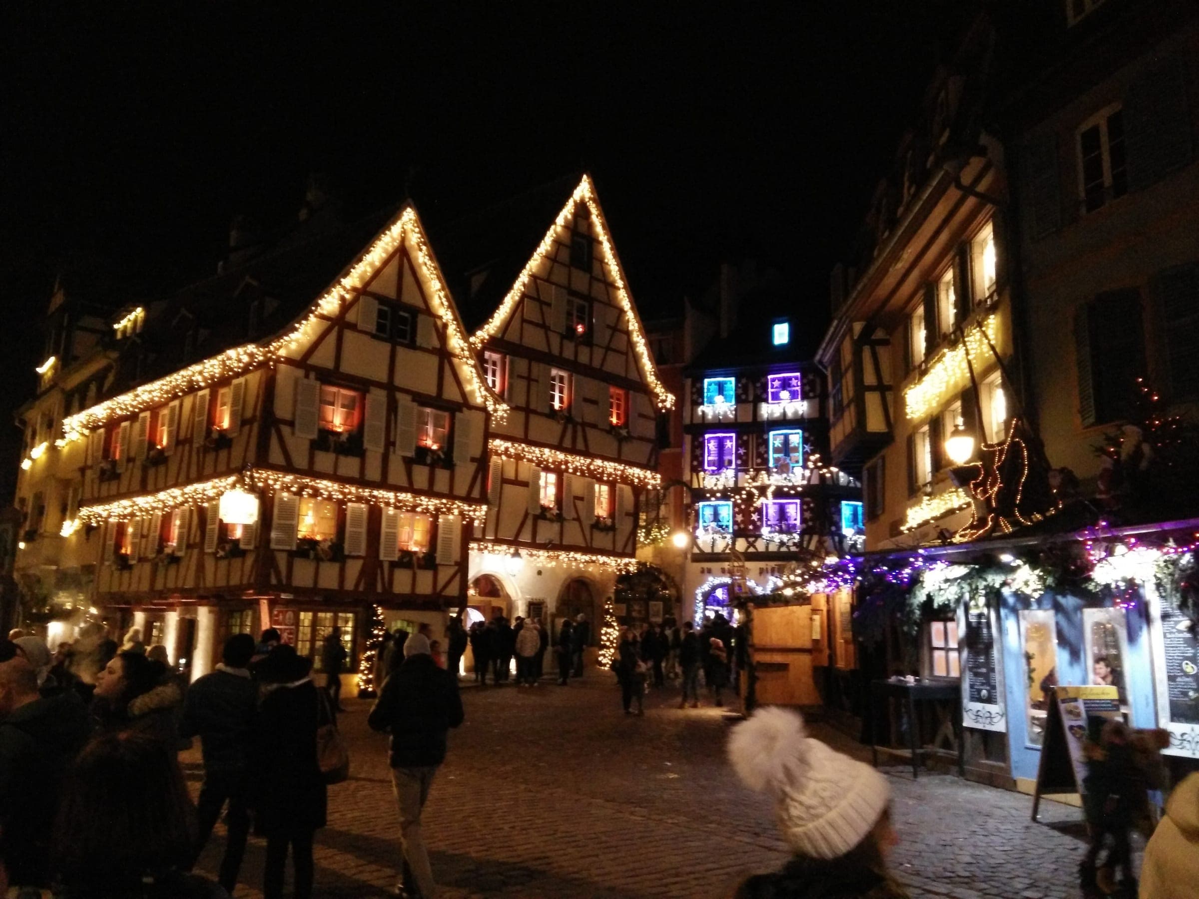 Strings of lights trace the steep gables of half-timbered houses in Colmar as people walk below at night.