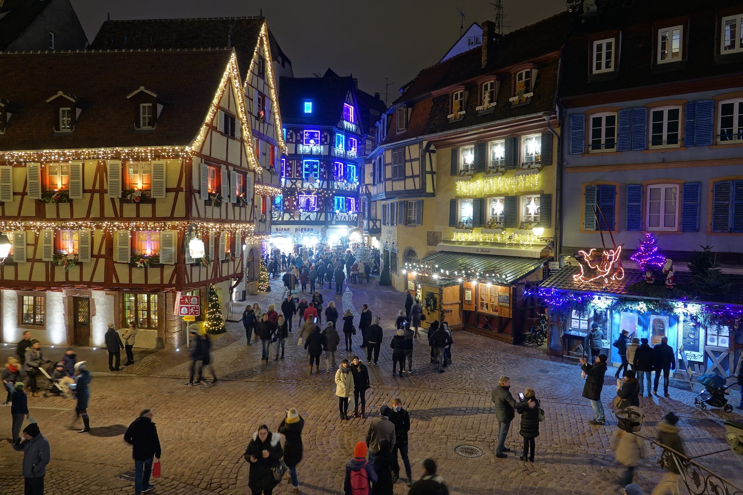 Crowds cross the square below the Koïfhus in Colmar while lights outline the gables of the old houses.