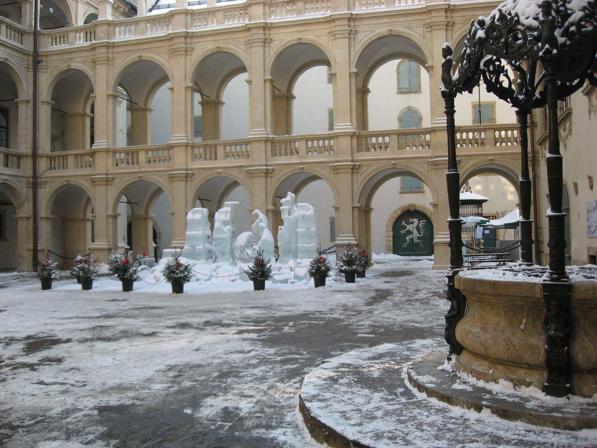 Snow covers the arcaded Landhaushof in Graz, with the ice nativity standing beyond the bronze well canopy.