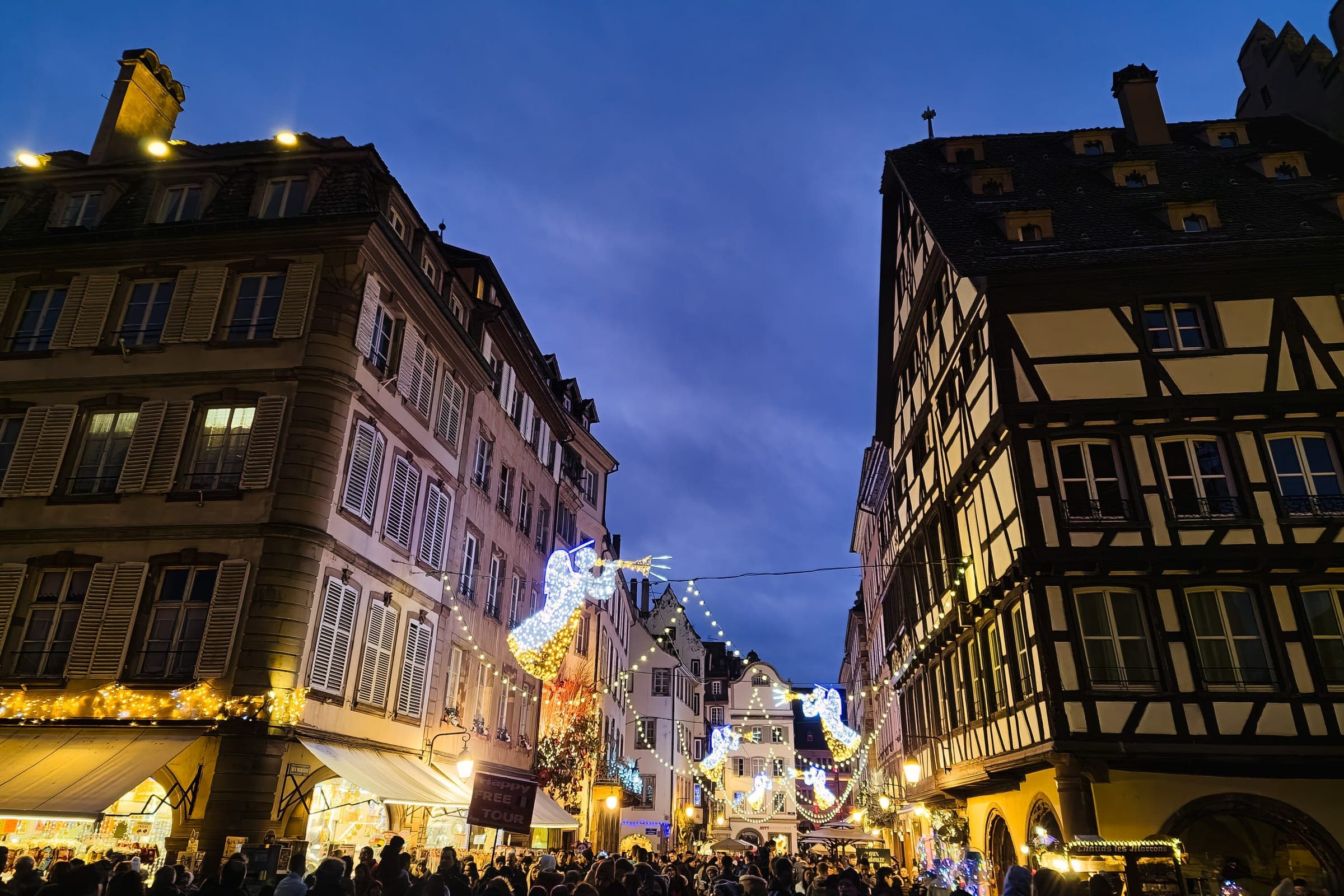 Lit angels and garlands cross a street of tall half-timbered houses in Strasbourg at dusk.
