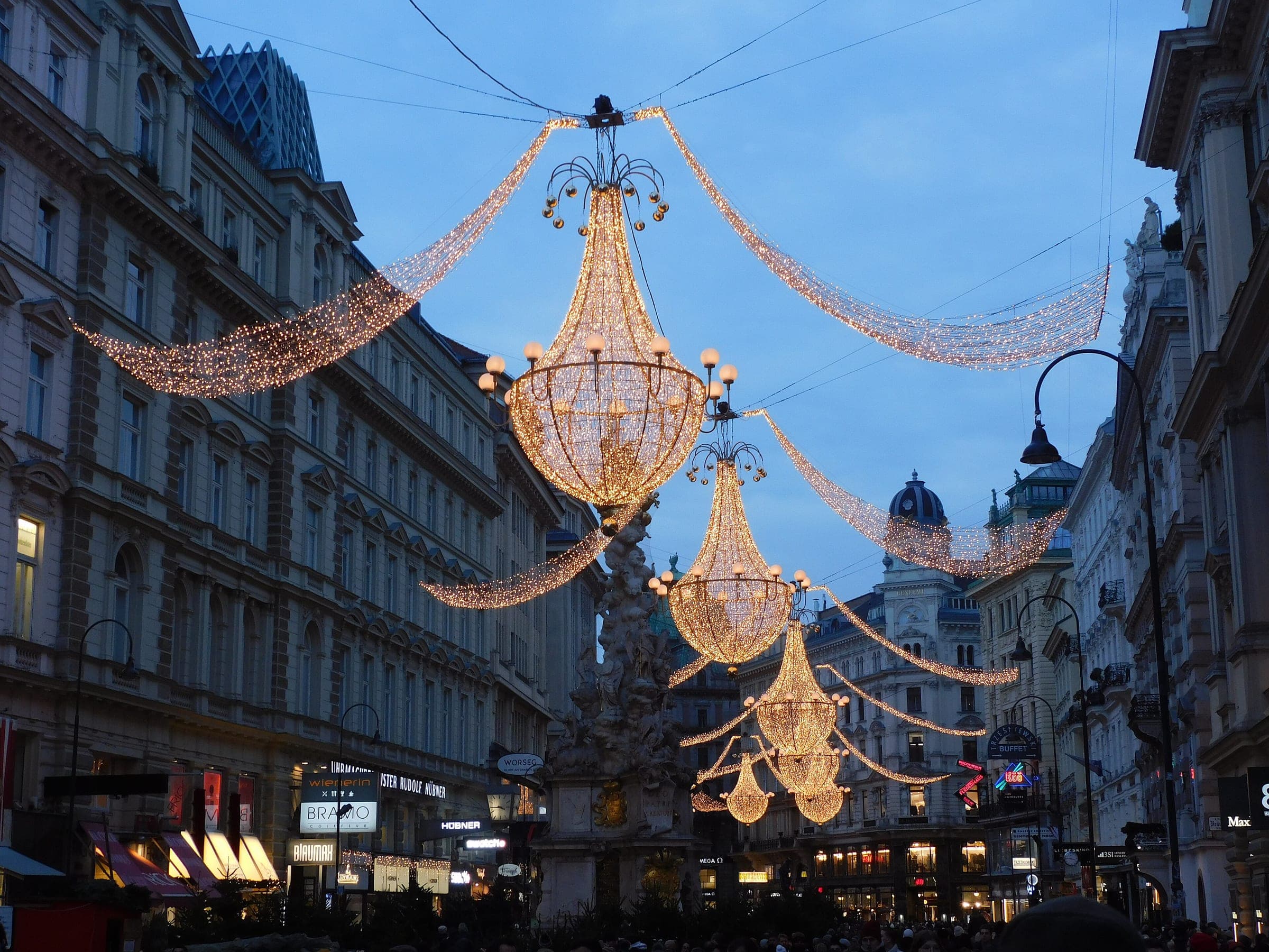 Chandelier-shaped Christmas lights hang over the Graben in Vienna at dusk, with the Plague Column below them.