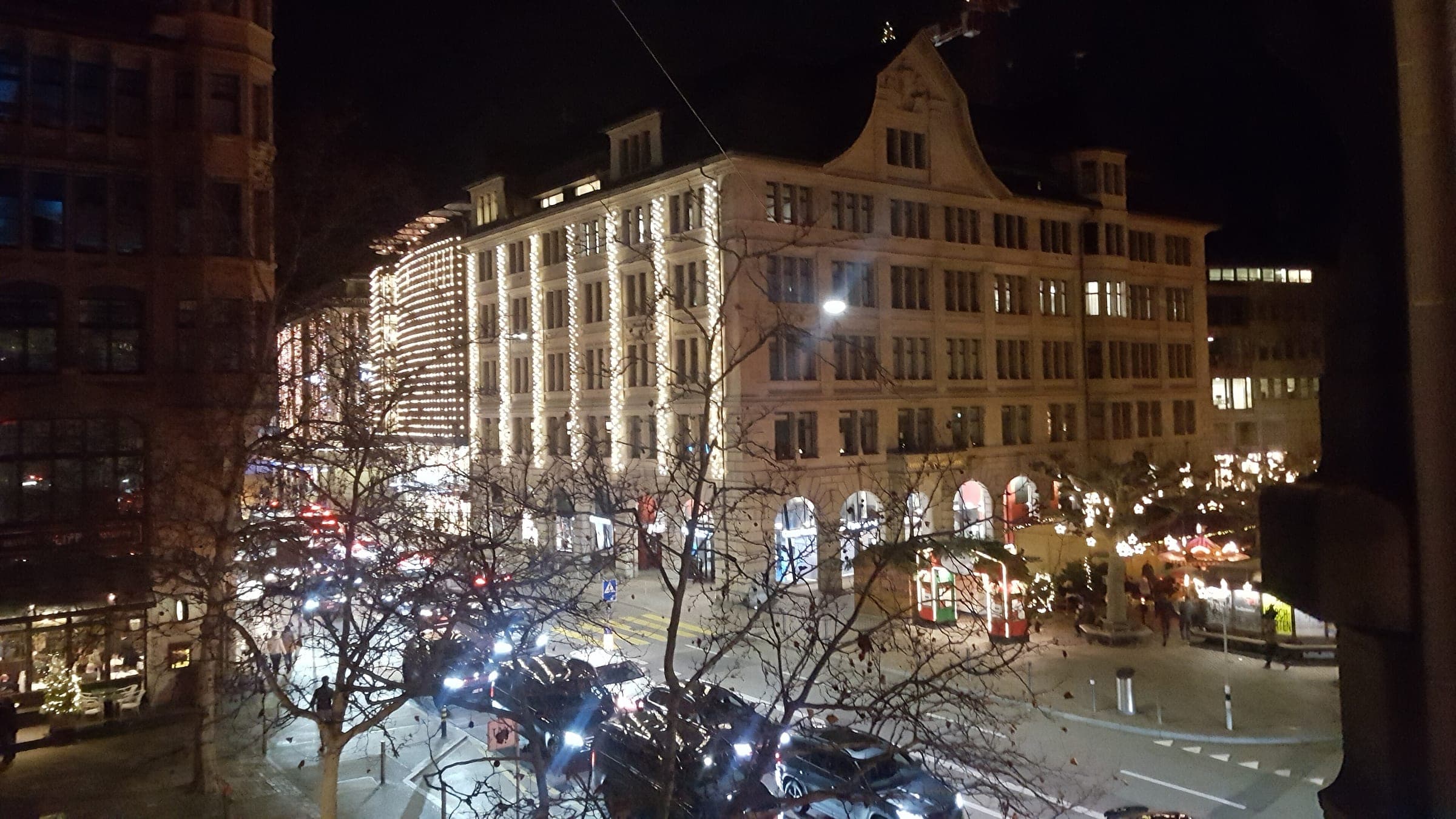 Lit department store facades and bare trees line a busy Zurich street on a December night