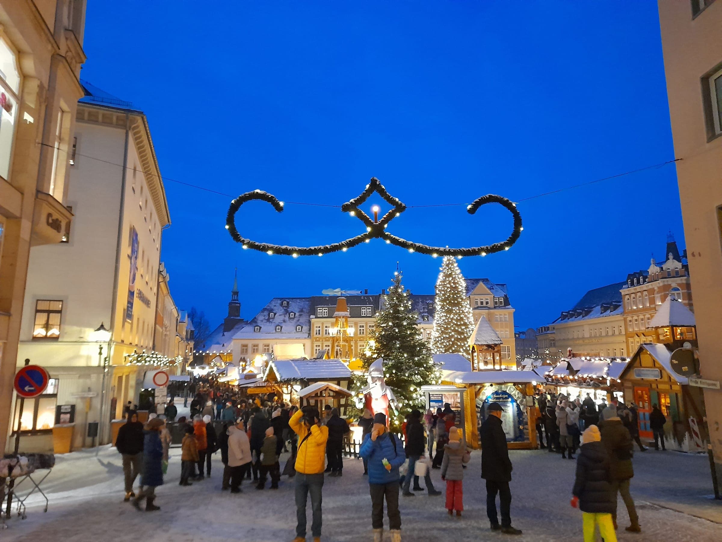 Crowds stroll under a lit garland toward the snowy stalls and trees of the Annaberg-Buchholz market at blue hour.