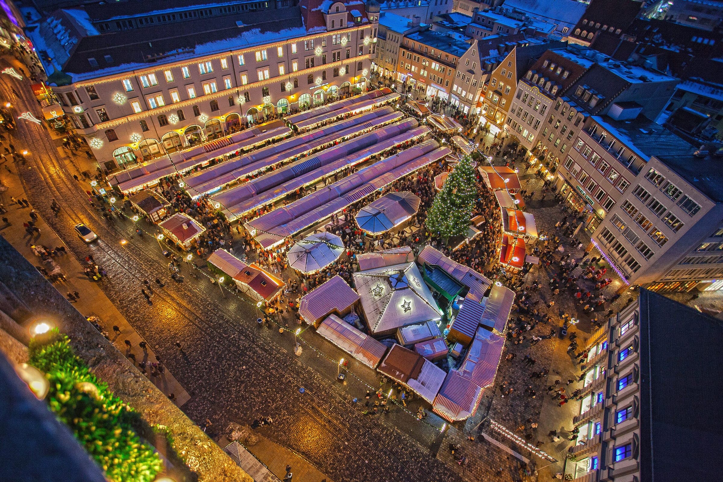 Lit stall roofs of the Augsburg Christkindlesmarkt fill Rathausplatz at night, seen from the Perlach Tower.