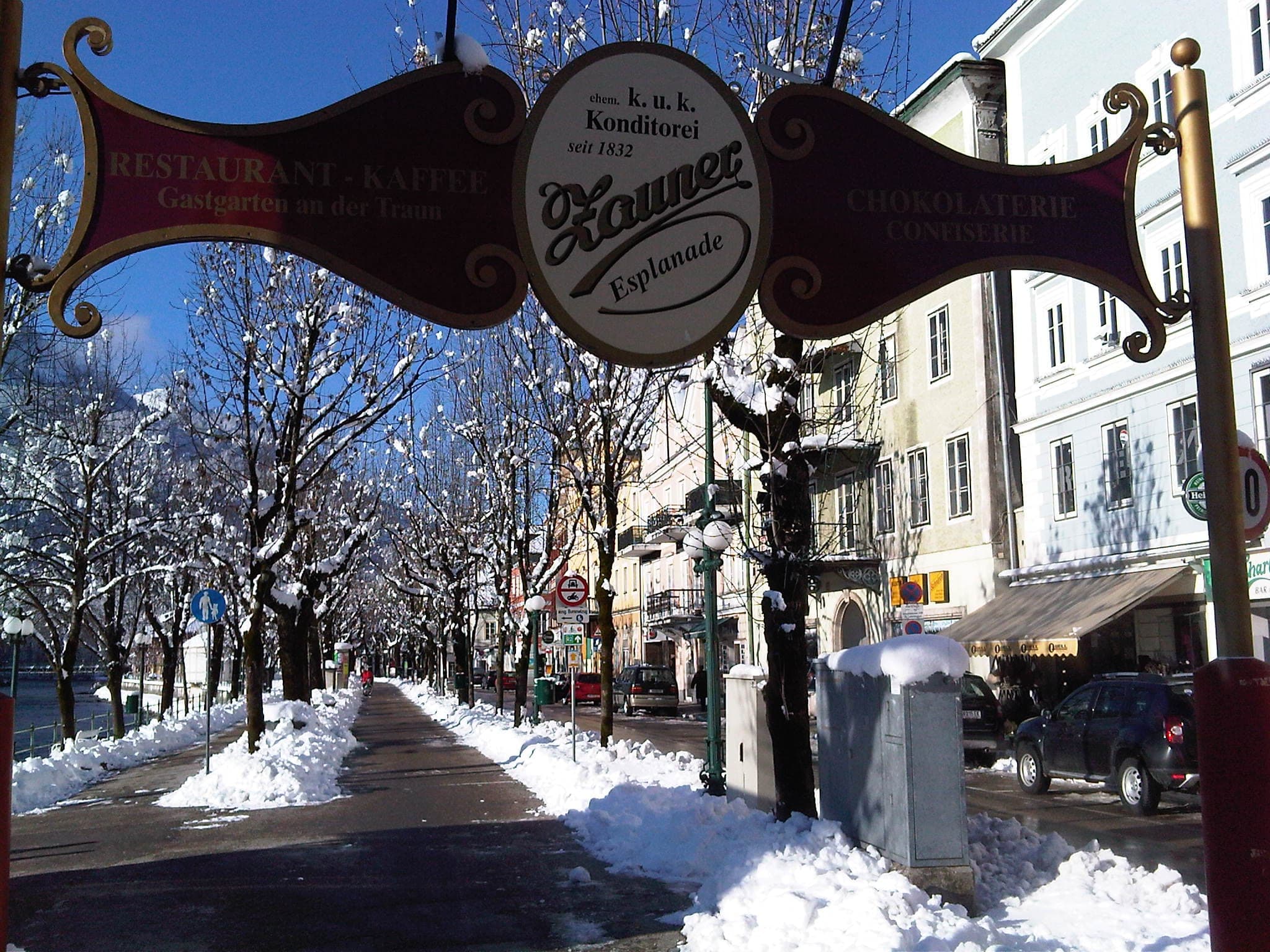 The Zauner Esplanade sign hangs over a snowy tree-lined promenade by the Traun in Bad Ischl