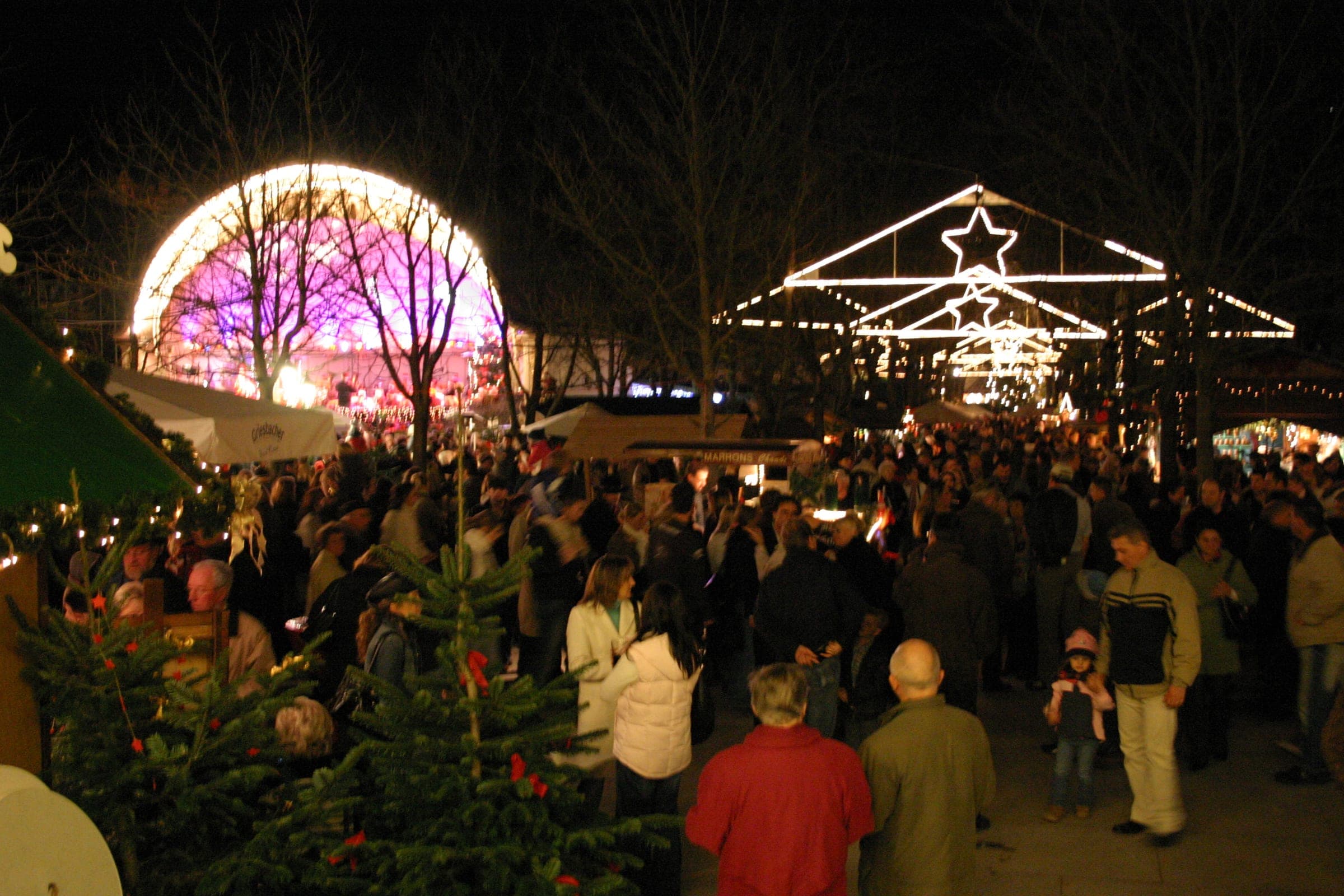Evening crowds fill the Christkindelsmarkt in Baden-Baden's Kurgarten under bare trees and star-shaped lights.