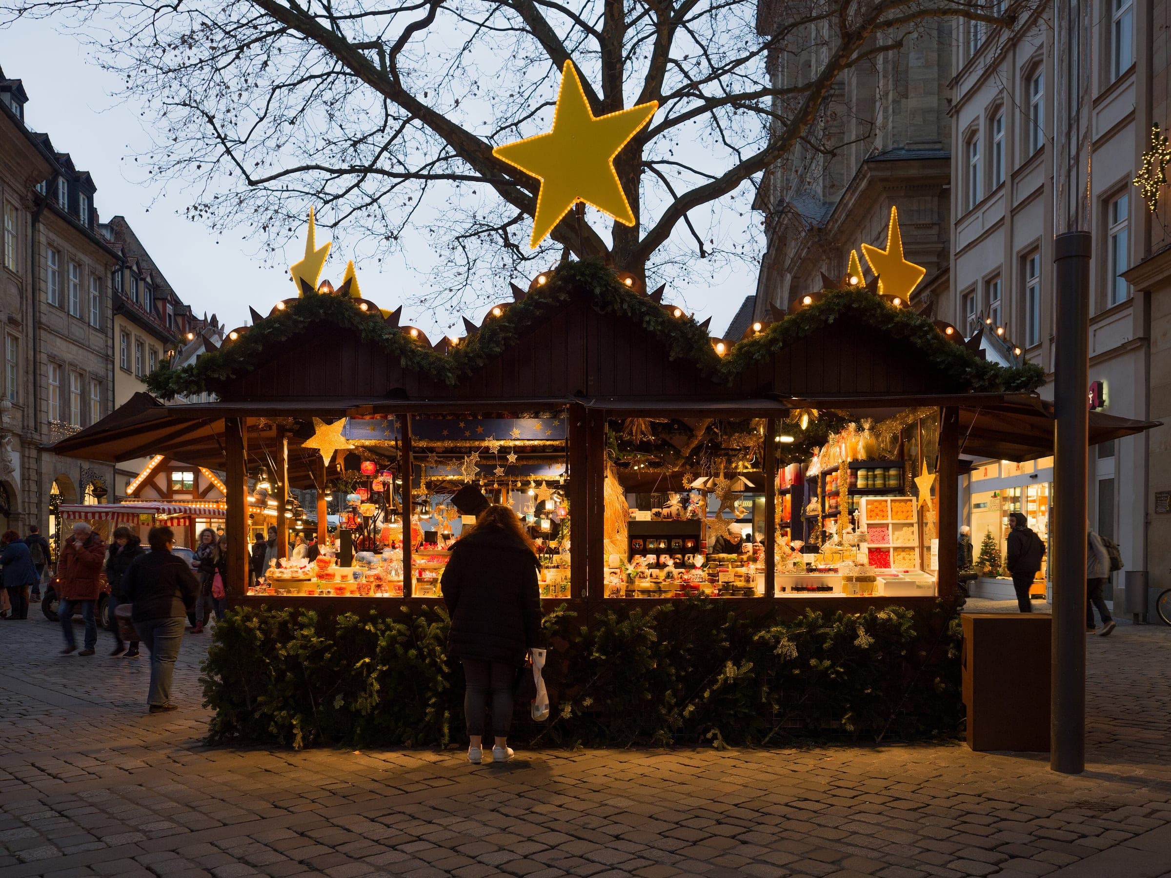 A shopper stands at a lit wooden stall under a gold star at the Bamberg Christmas market at dusk