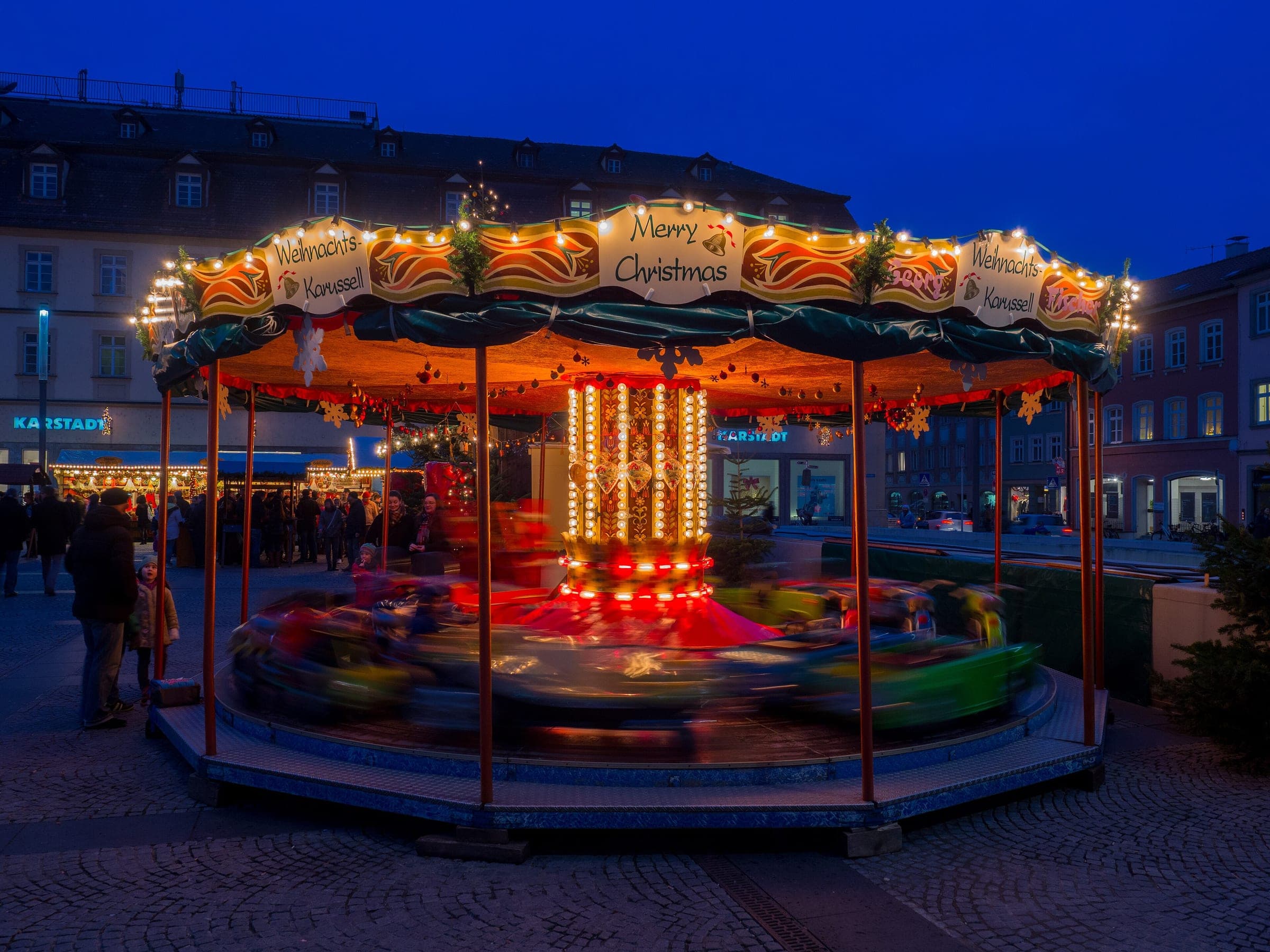 A merry-go-round spins under garlands at the Bamberg Christmas market on Maximiliansplatz at blue hour