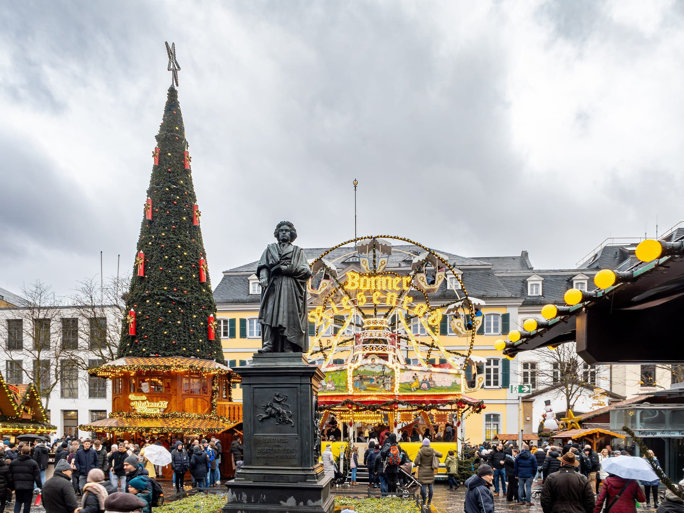 The Beethoven statue stands between a tall decorated tree and a carousel on Bonn's Münsterplatz