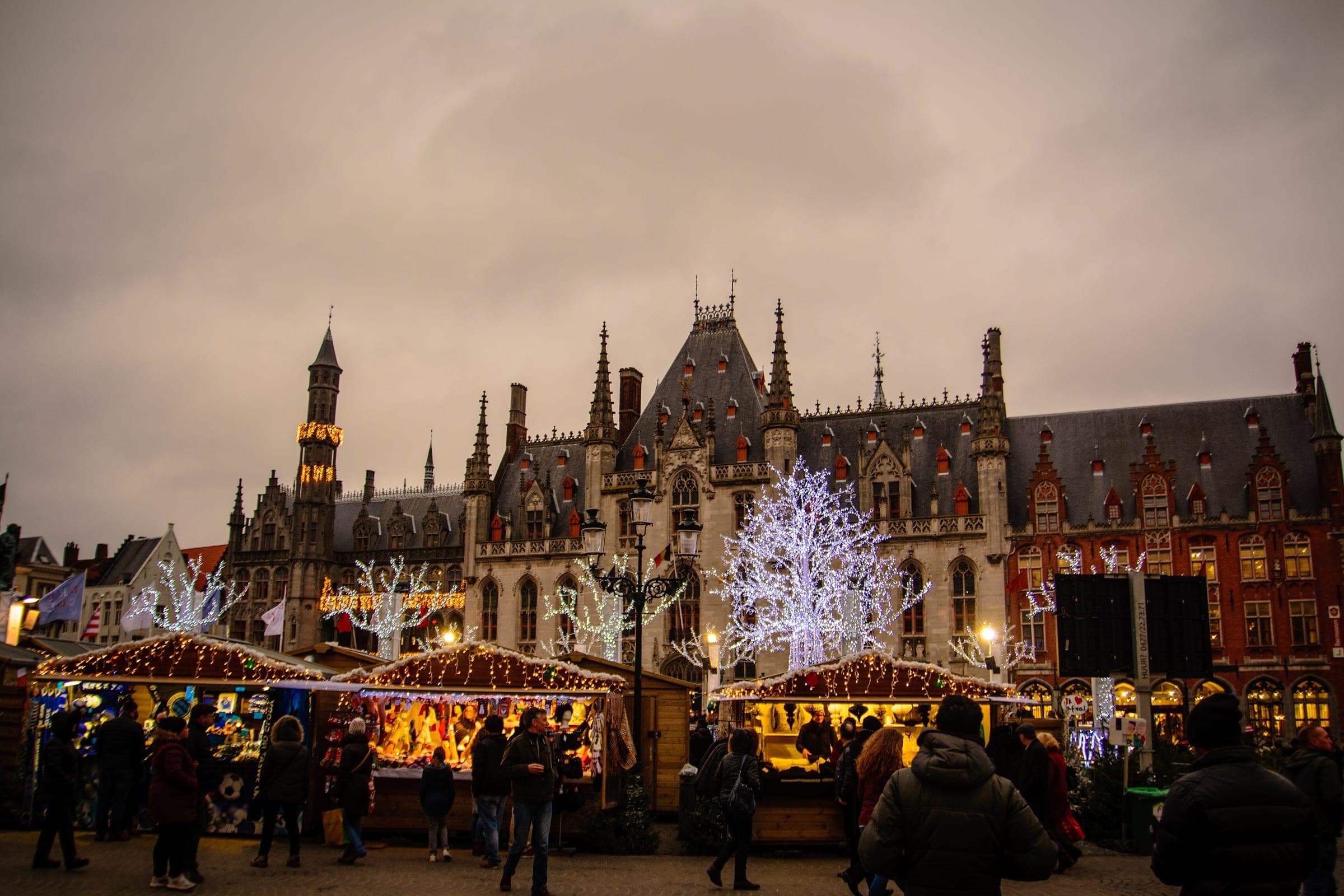 Lit chalets fill the Markt in Bruges below the Provinciaal Hof and its white light trees at dusk.