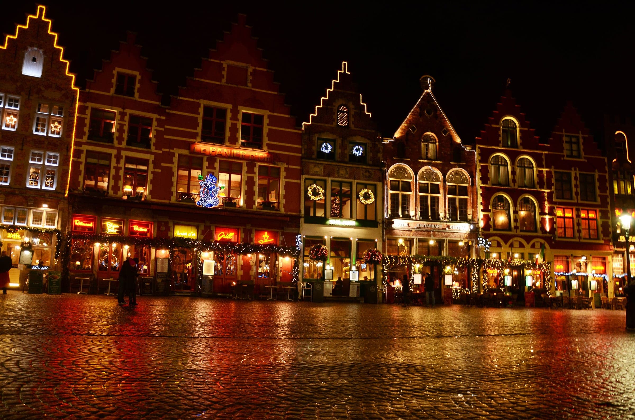 Wreaths and string lights hang on the guild houses of the Markt in Bruges on a wet winter night.