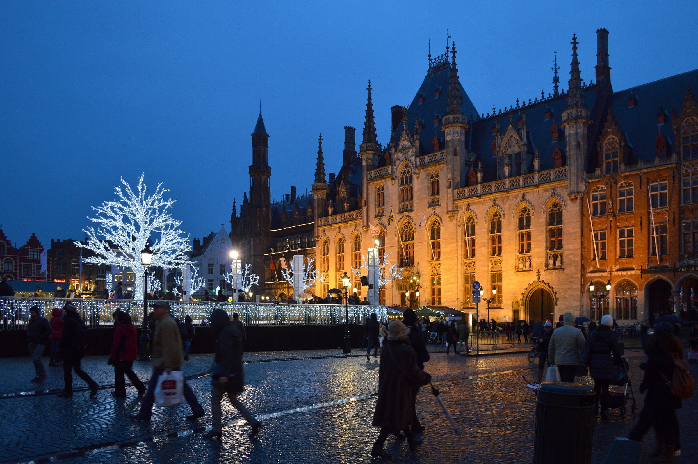 Shoppers cross the Markt in Bruges at blue hour as the Provinciaal Hof and a white light tree glow behind the rink.