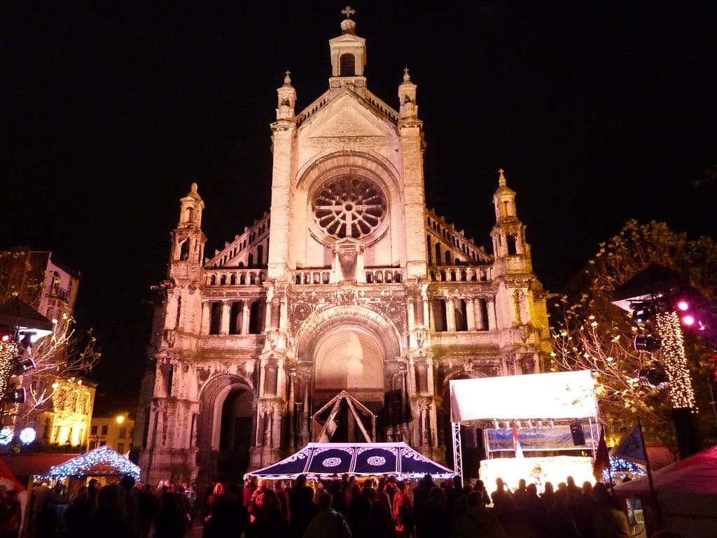 Floodlit Sainte-Catherine church rises over market stalls and a crowd on its square in Brussels at night.