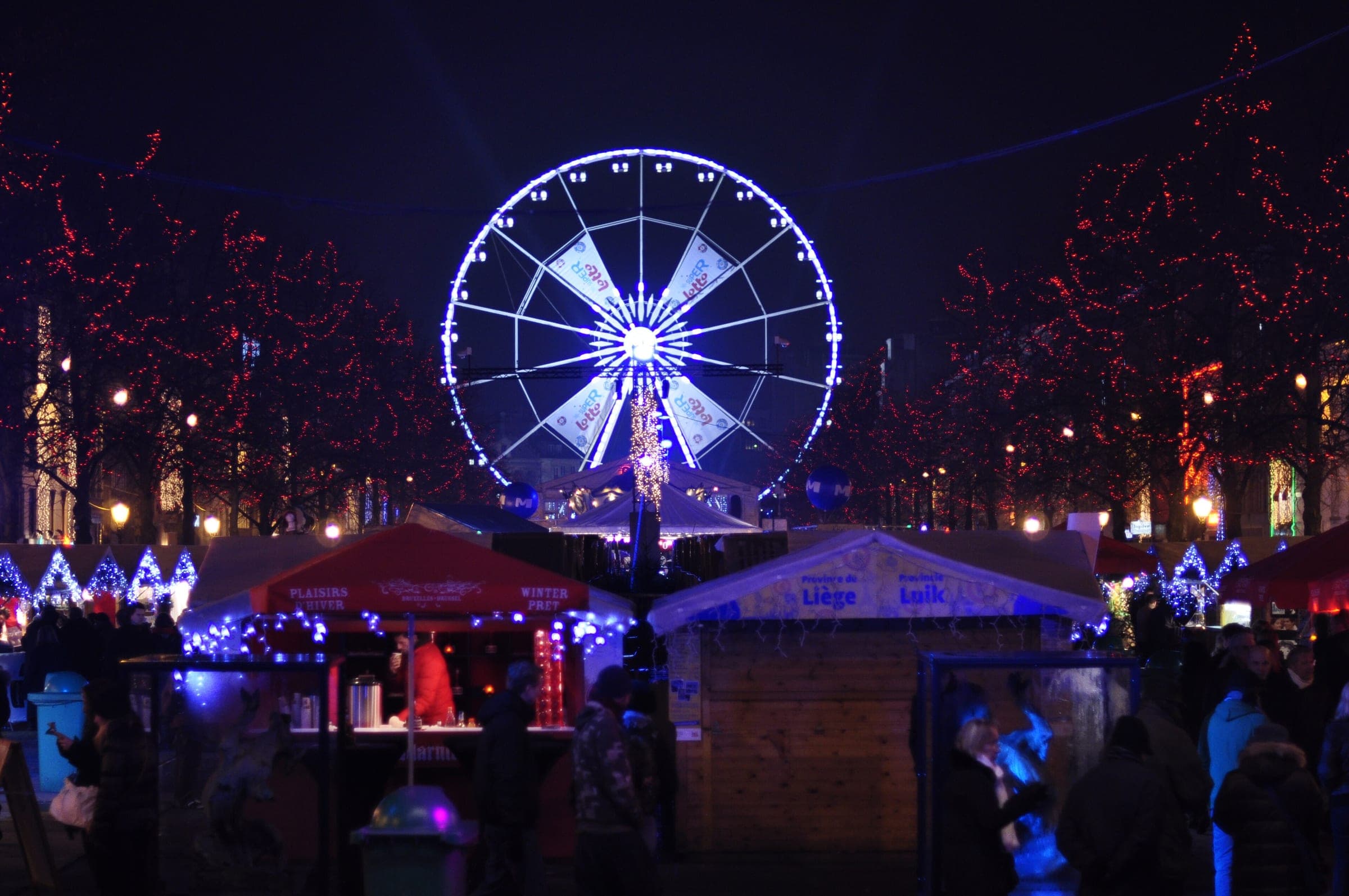 The Ferris wheel glows white above market chalets and red-lit trees at Winter Wonders in Brussels.