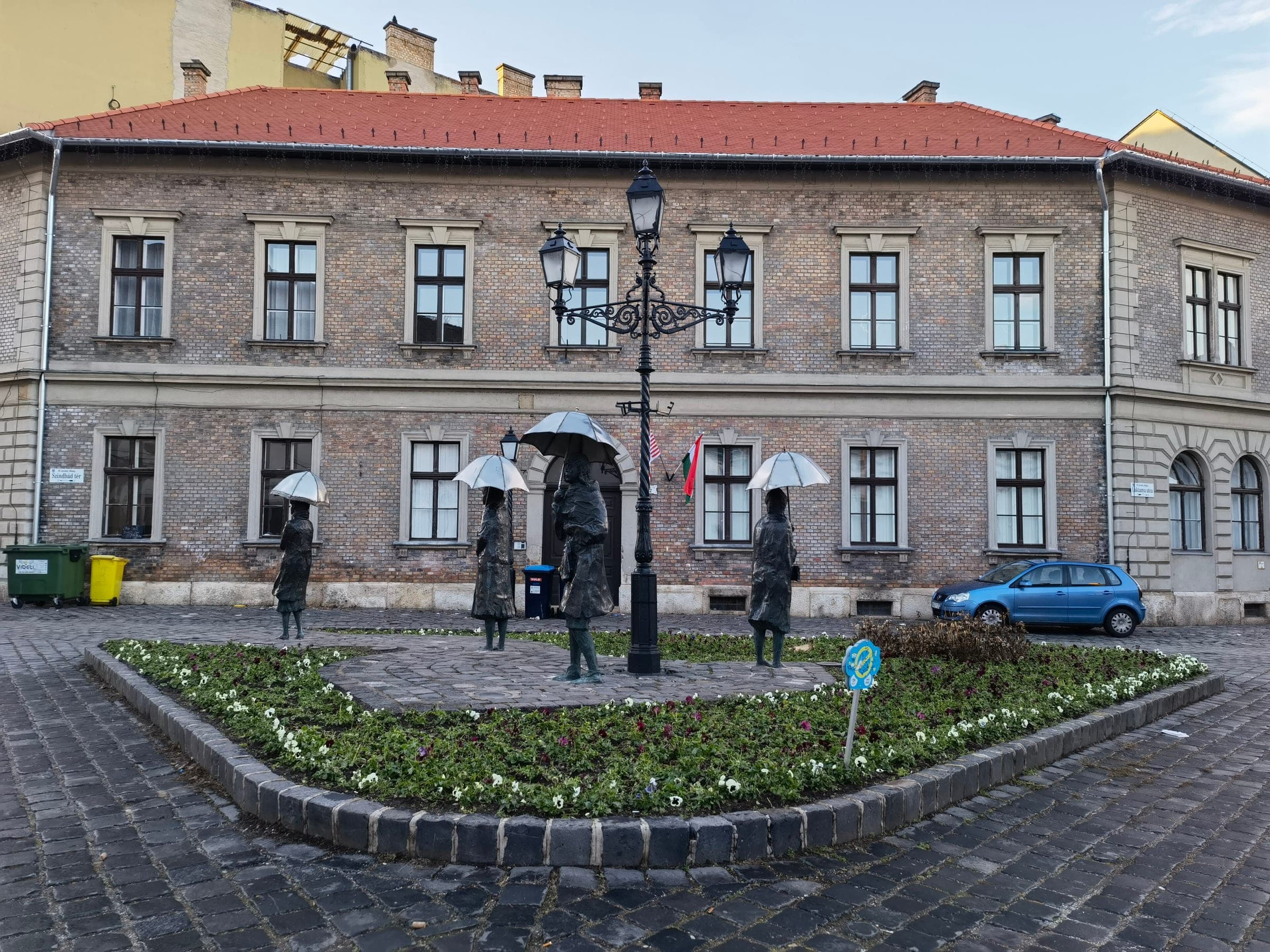Bronze women holding umbrellas stand in a flowerbed on Fő tér in Óbuda, Budapest, in December.