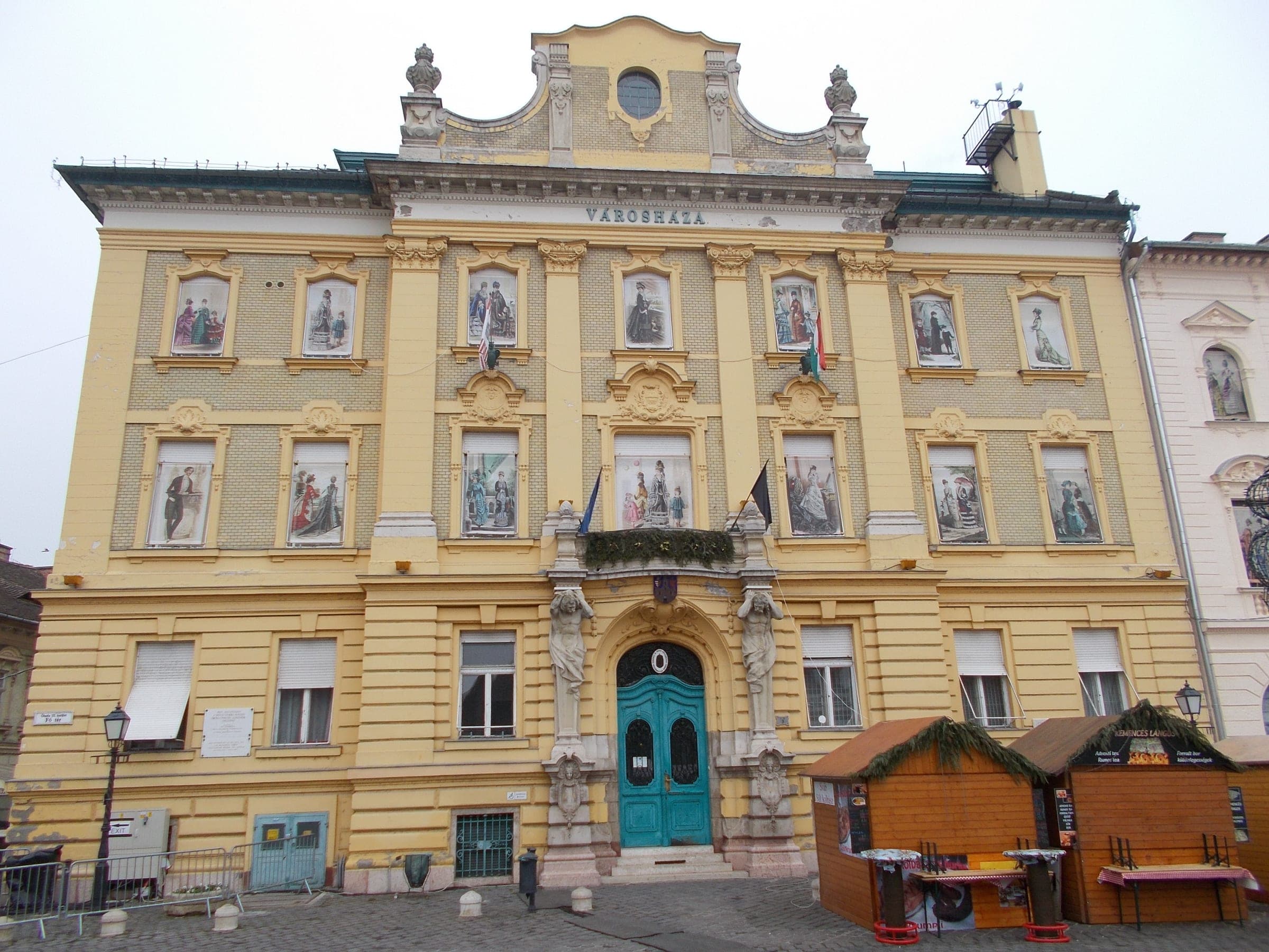 Wooden market huts with fir garlands stand beside the yellow Óbuda Town Hall on Fő tér in Budapest.