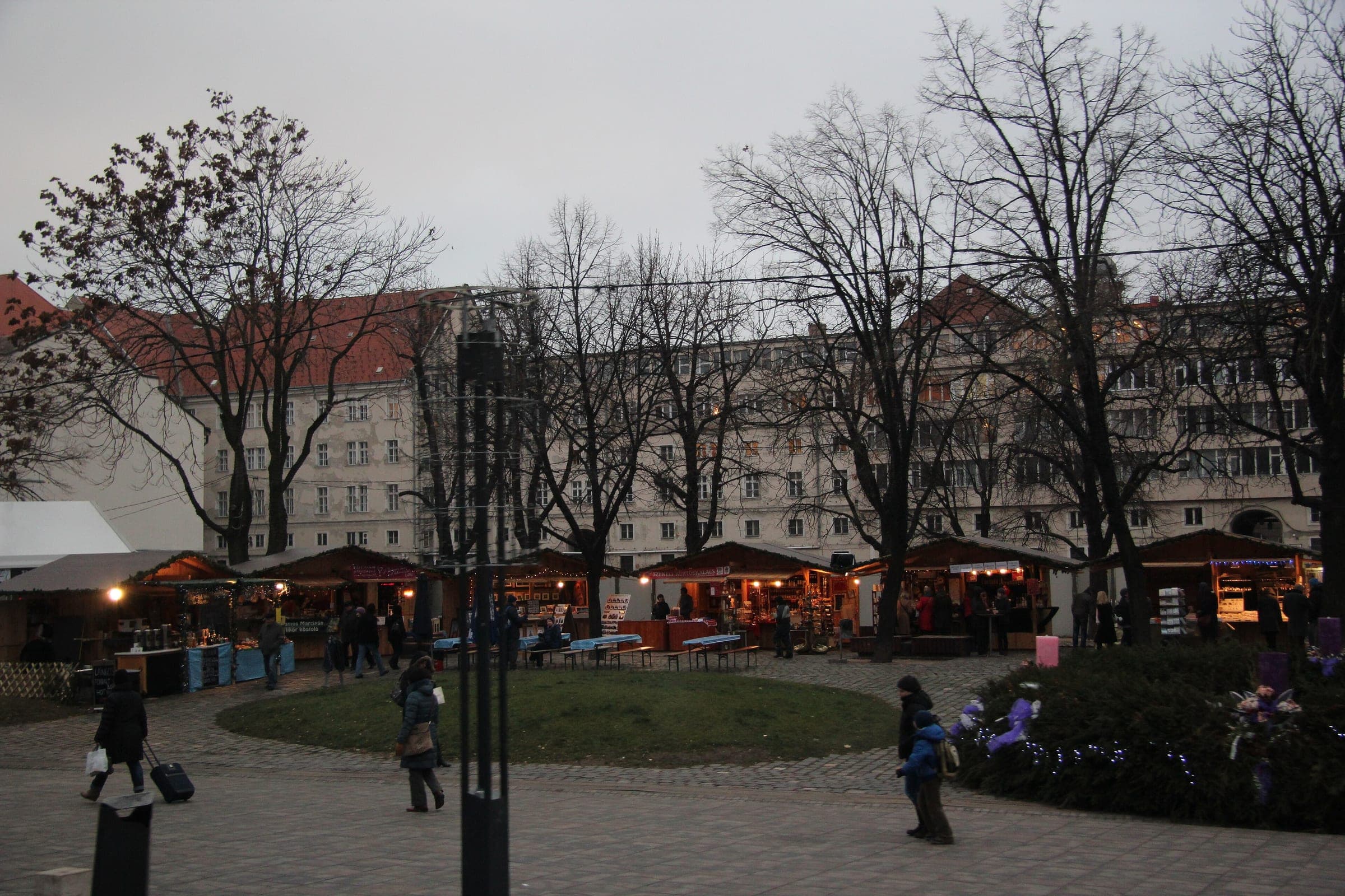 Market stalls glow under bare plane trees in Városháza Park in Budapest on a grey December evening.