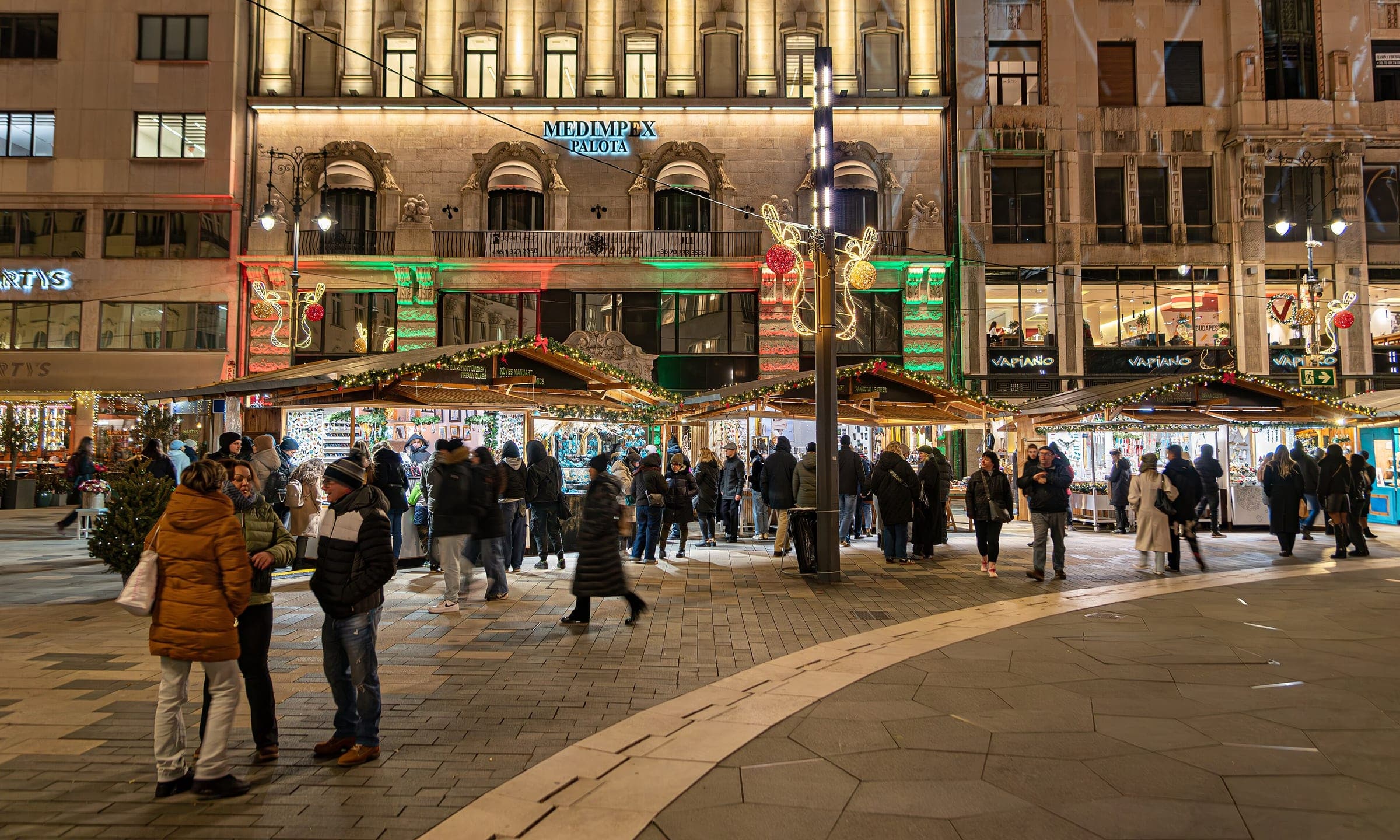 Shoppers pass a row of wooden stalls in front of the lit Medimpex house on Vörösmarty tér in Budapest.