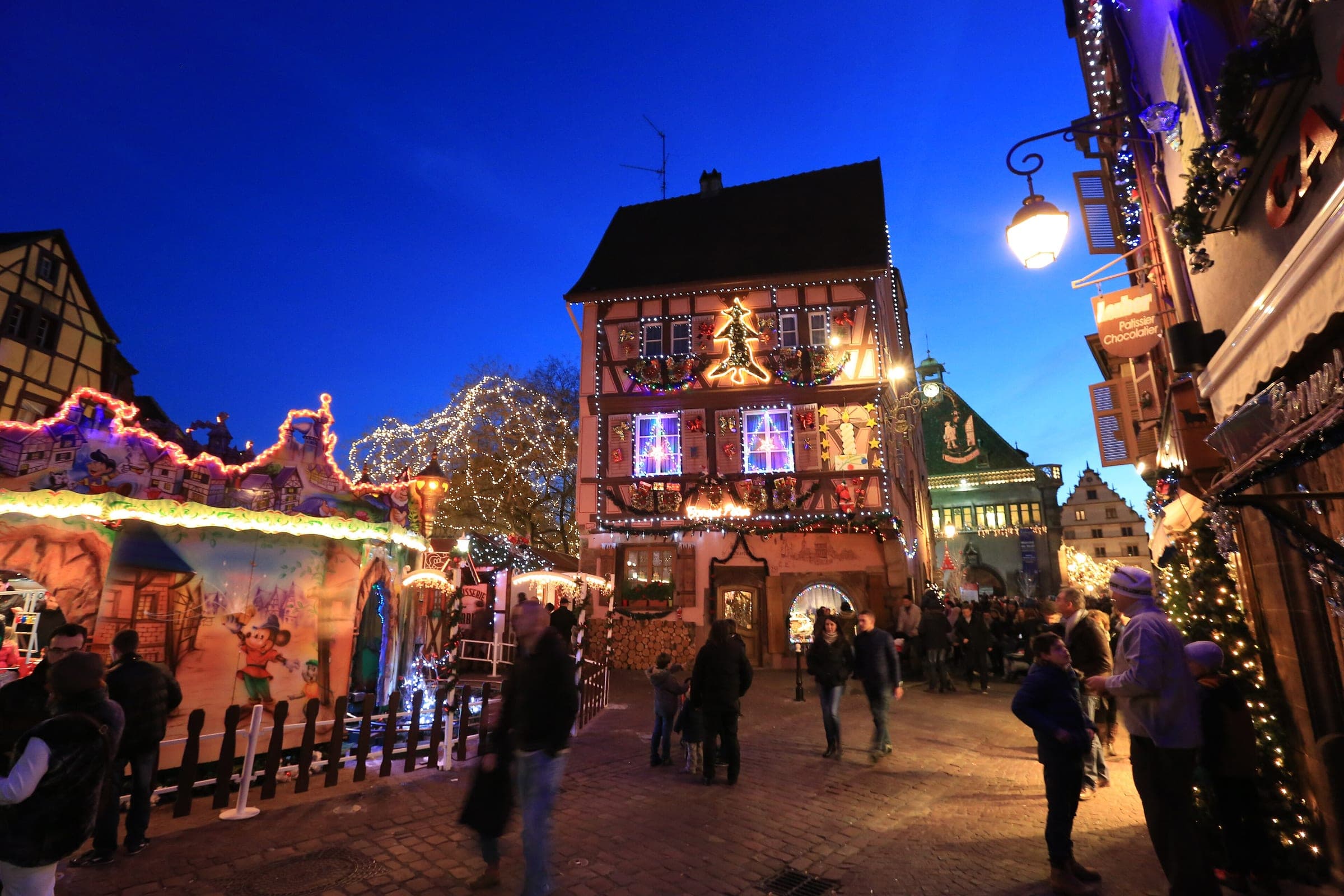 Visitors stroll past a carousel and the lit Maison du Pèlerin beside the Ancienne Douane market in Colmar at dusk.