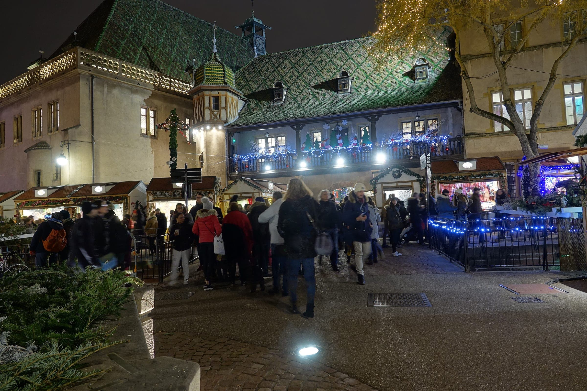 Crowds fill Place de l'Ancienne Douane in Colmar in front of the Koïfhus and its row of lit chalets at night.
