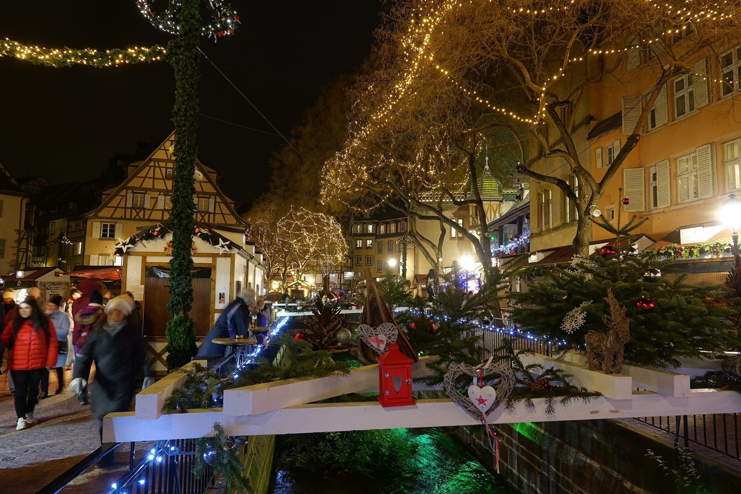 Lights hang in the trees over the Lauch as shoppers walk past the Ancienne Douane chalets in Colmar at night.