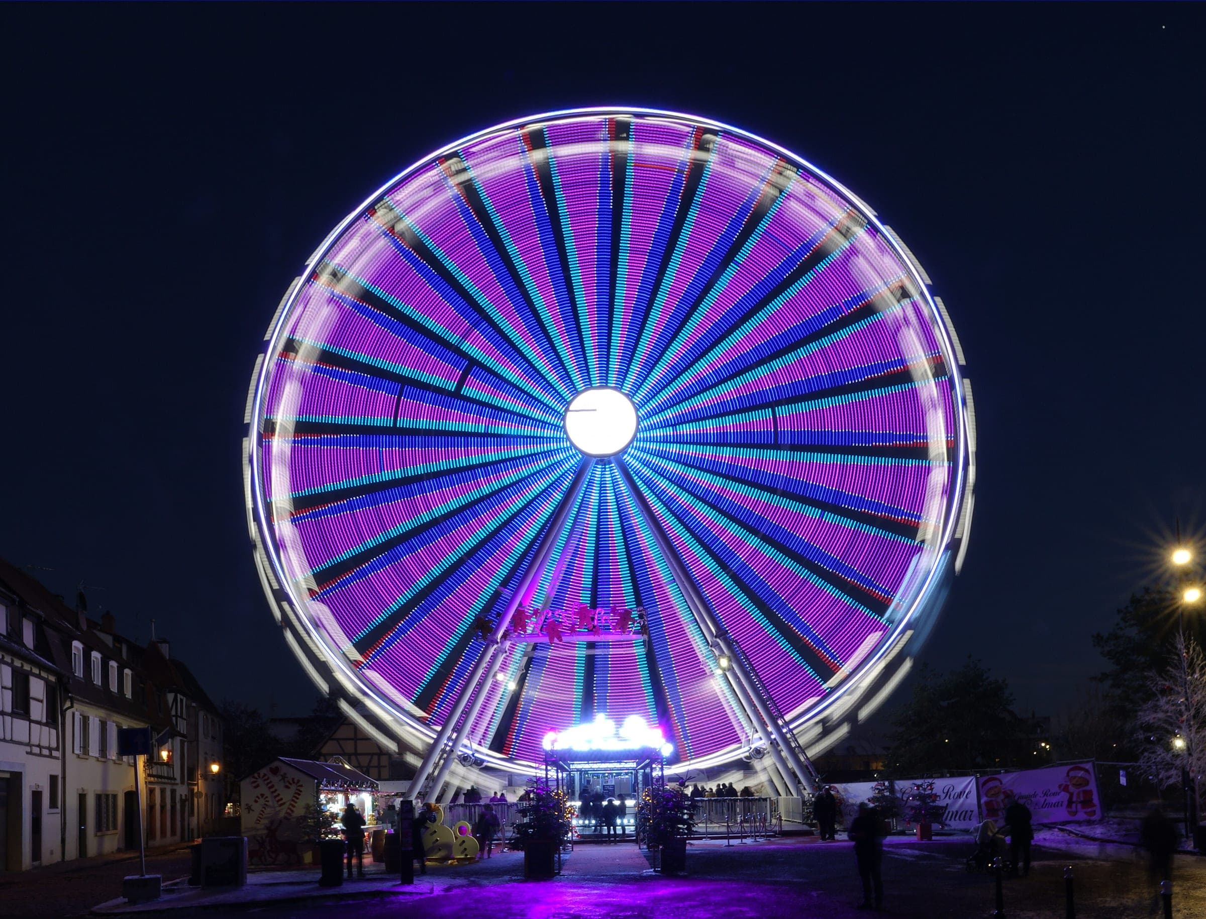 The Ferris wheel spins in purple light above the gourmet market chalets in Colmar on a December evening.