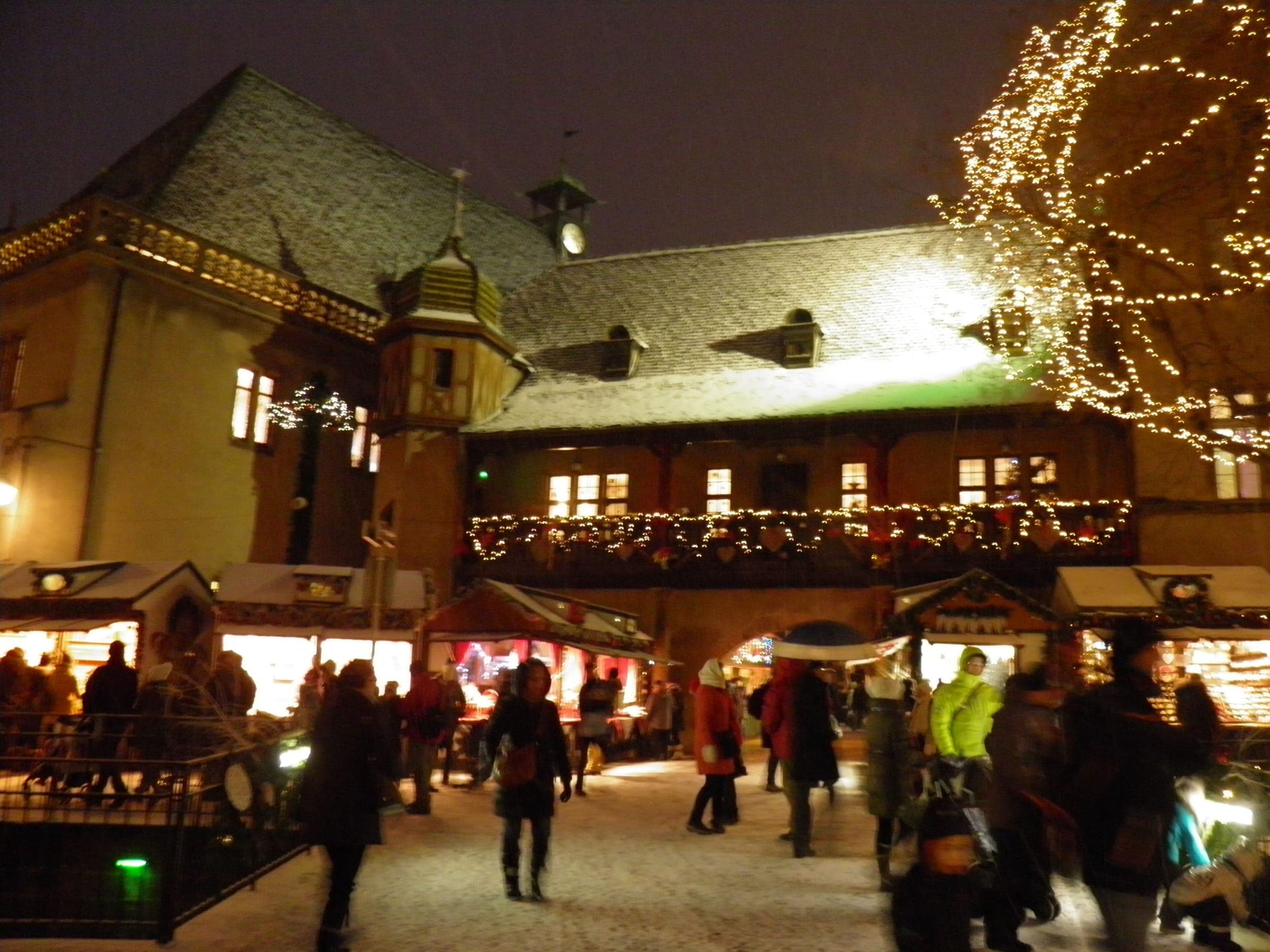 Lights trace the gallery and tiled roof of the Koïfhus in Colmar as crowds move past the market stalls at night.