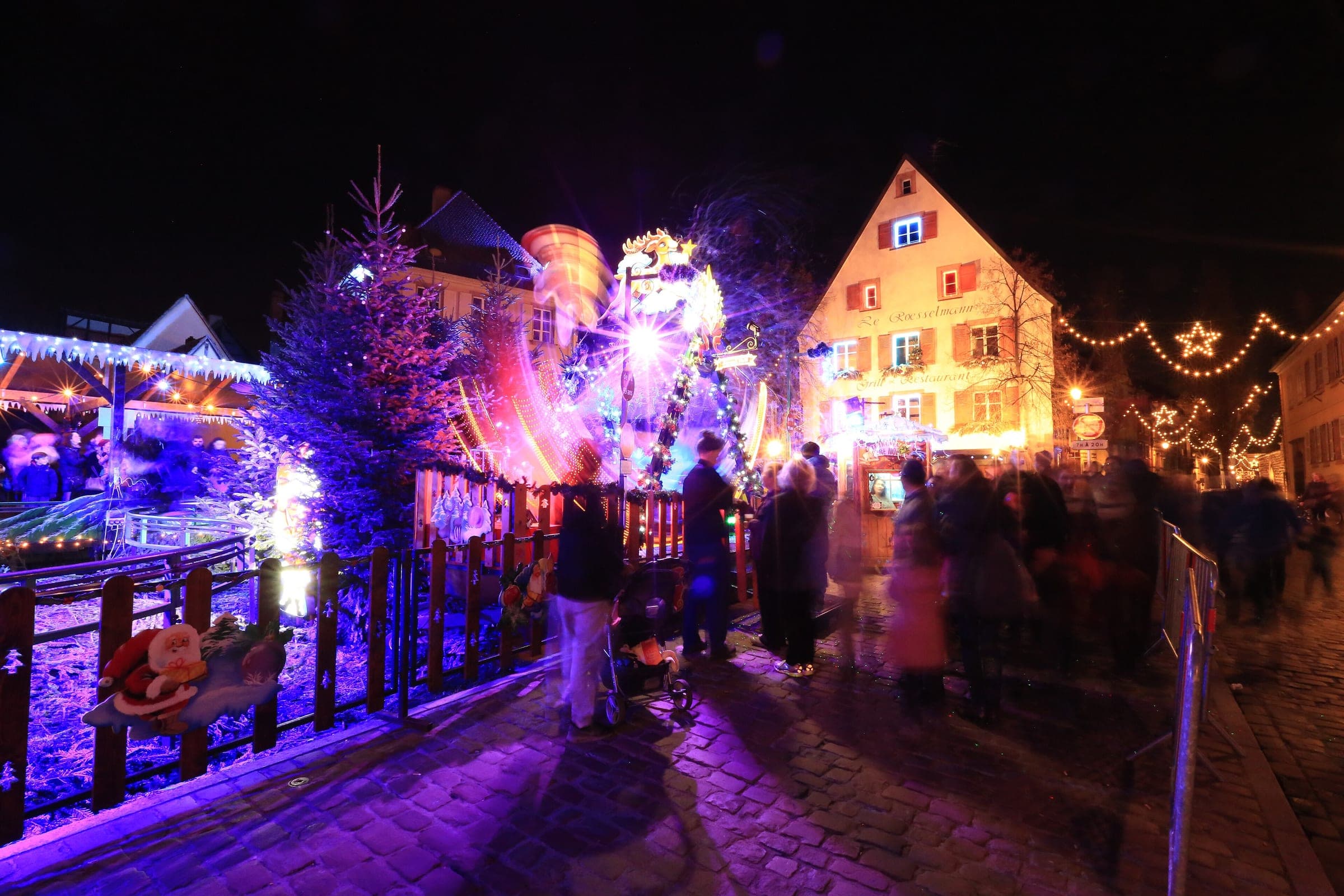 Purple and gold lights fill Place des Six Montagnes Noires in Petite Venise as families walk through at night.