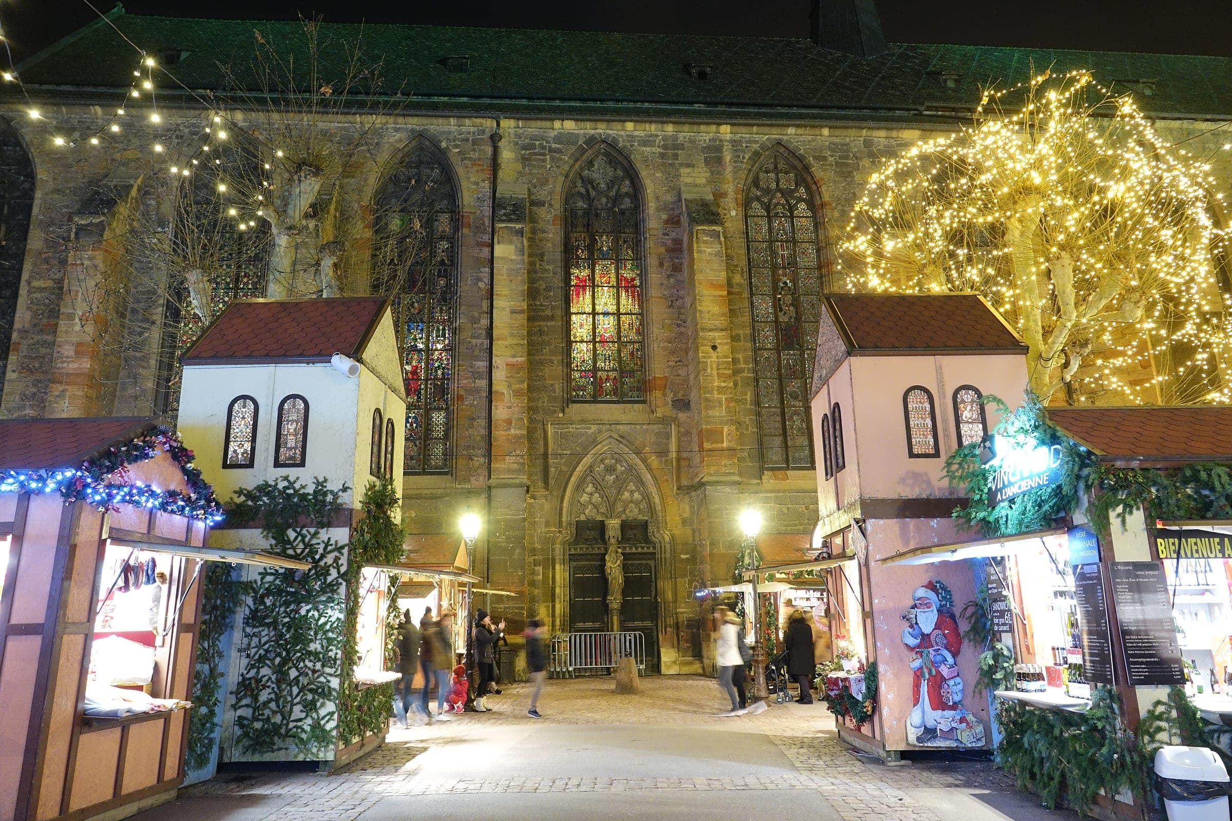 Chalets with pointed roofs stand in front of the lit Dominican church on Place des Dominicains in Colmar.