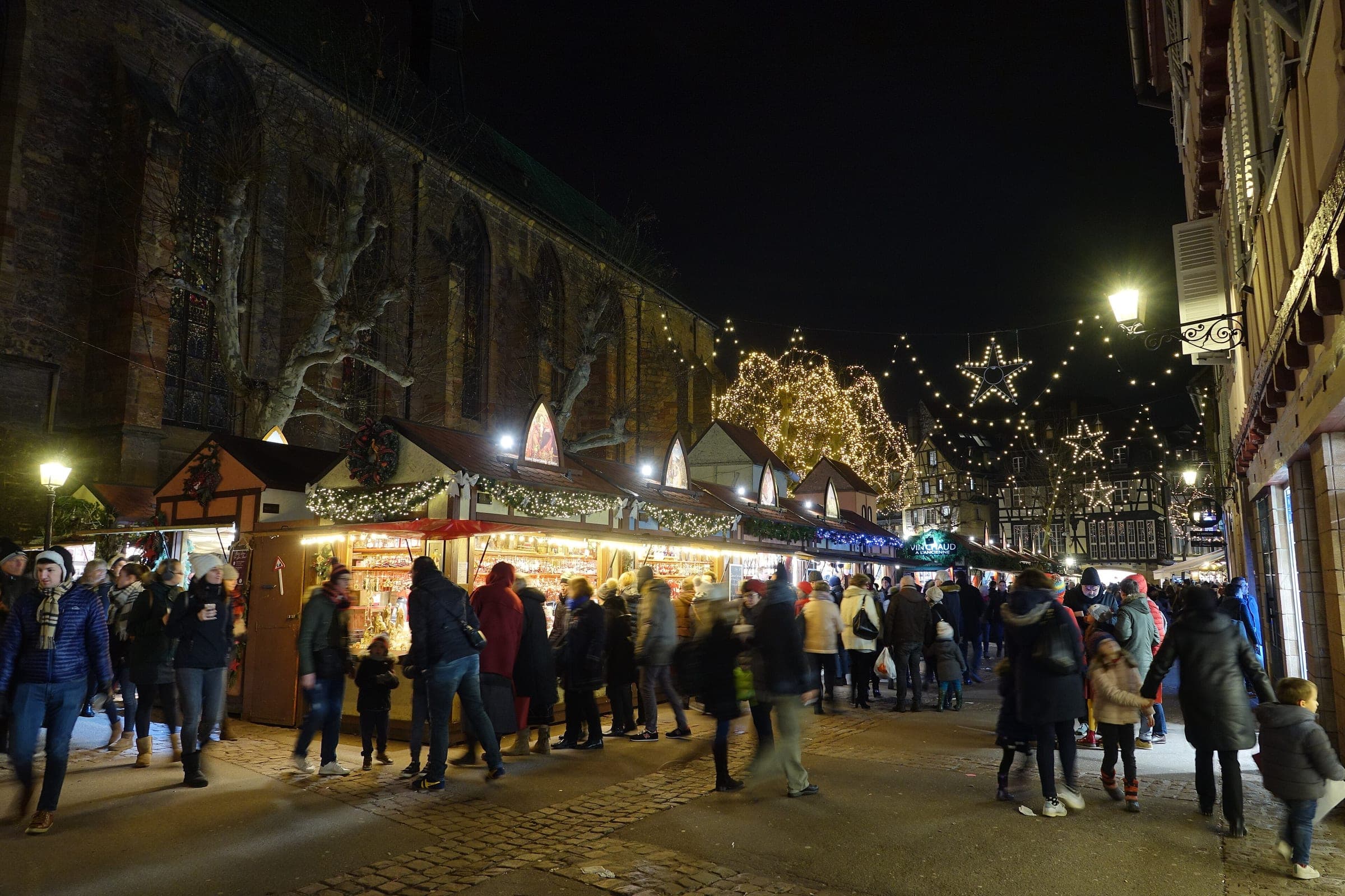 Shoppers walk between the lit chalets on Place des Dominicains in Colmar under strings of stars at night.