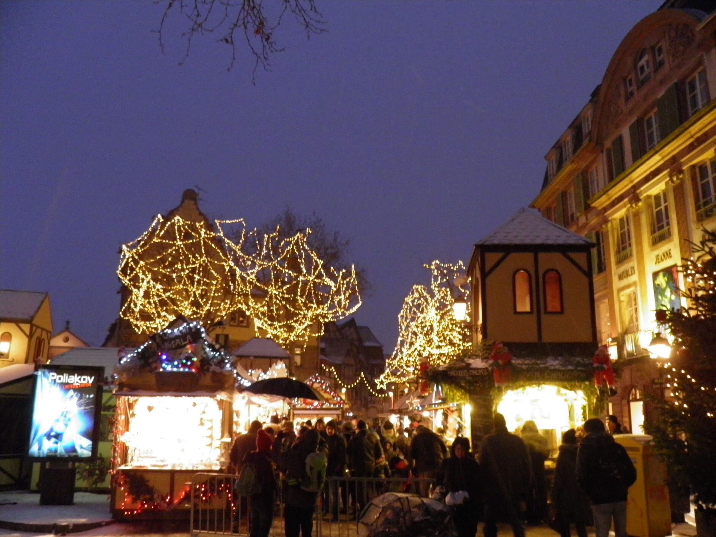 Lit trees and chalets glow under a violet evening sky on Place Jeanne d'Arc in Colmar as crowds gather.
