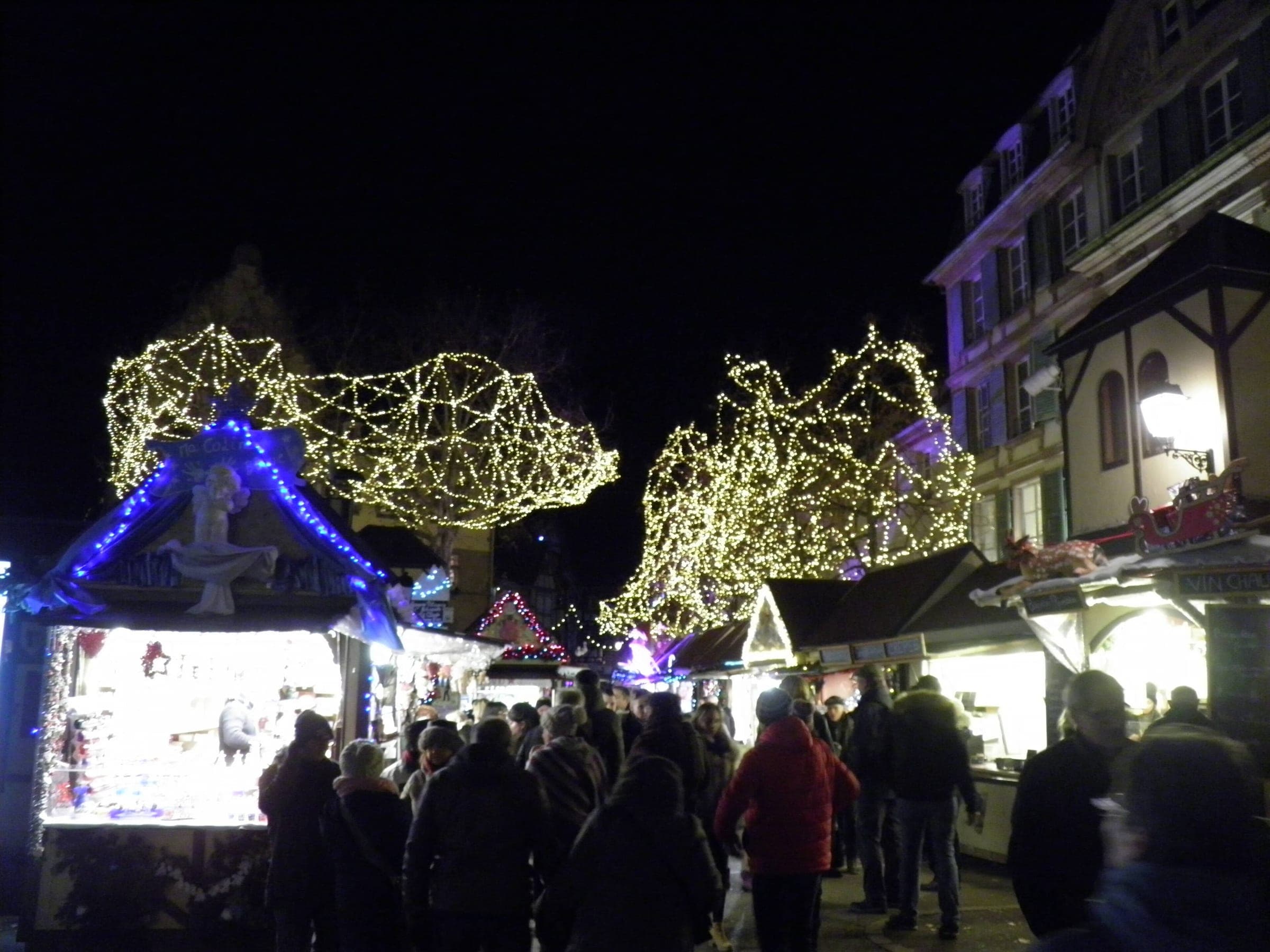 Visitors queue at the bright chalets of the Place Jeanne d'Arc market in Colmar under trees wrapped in lights.