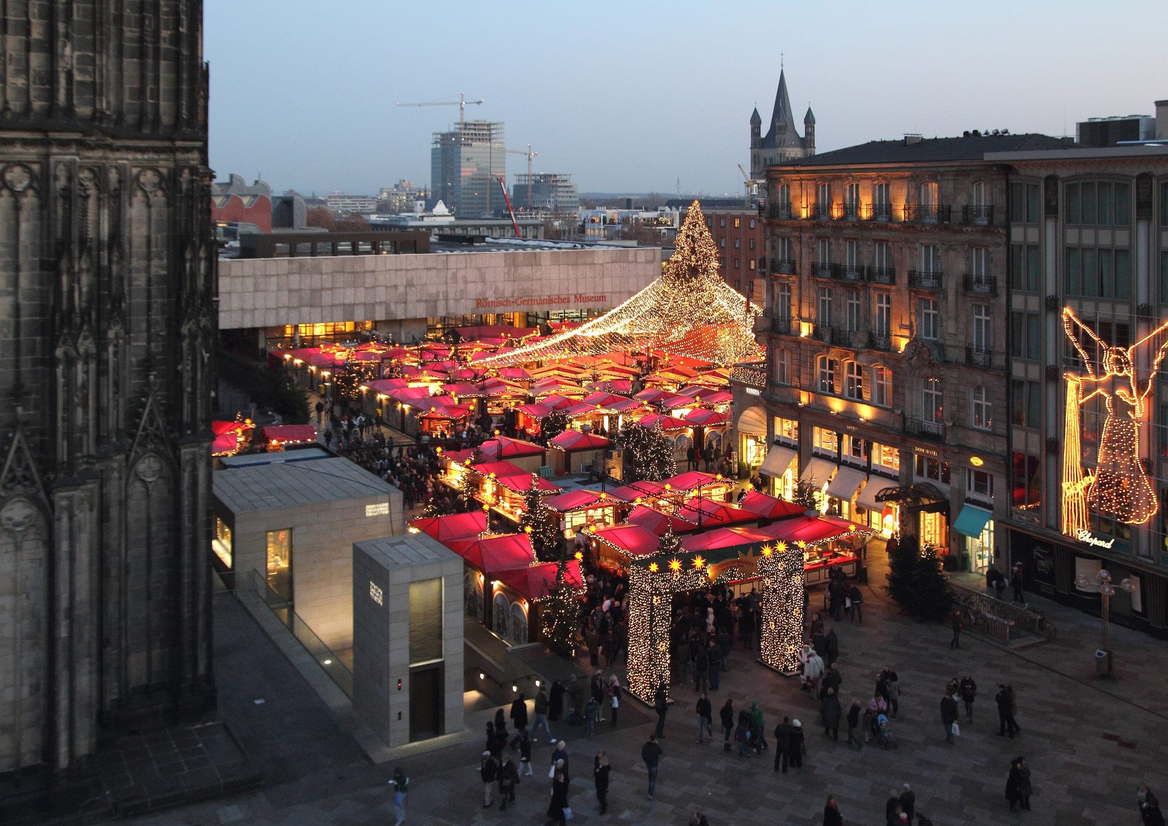 Red stall roofs of the Cologne Cathedral Christmas market fill Roncalliplatz at dusk beside the cathedral