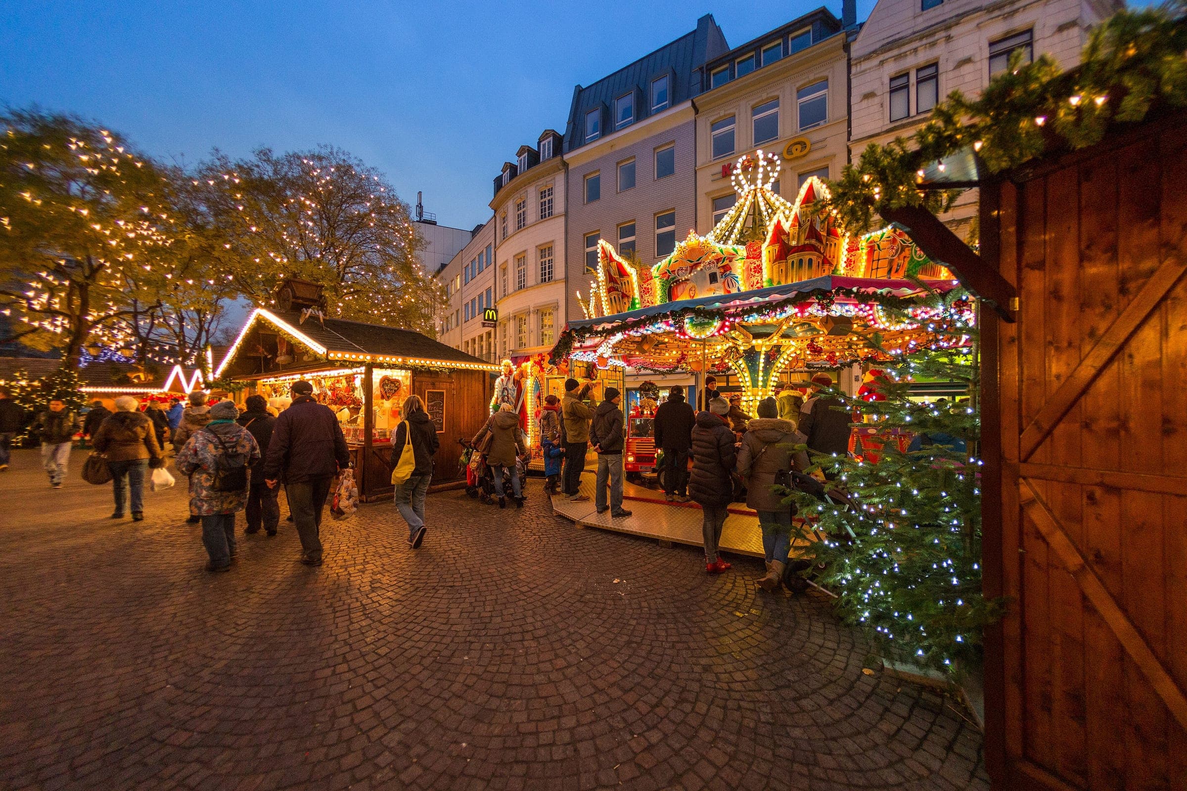 Market huts and a lit carousel stand on the cobbles of Cologne's Alter Markt at blue hour