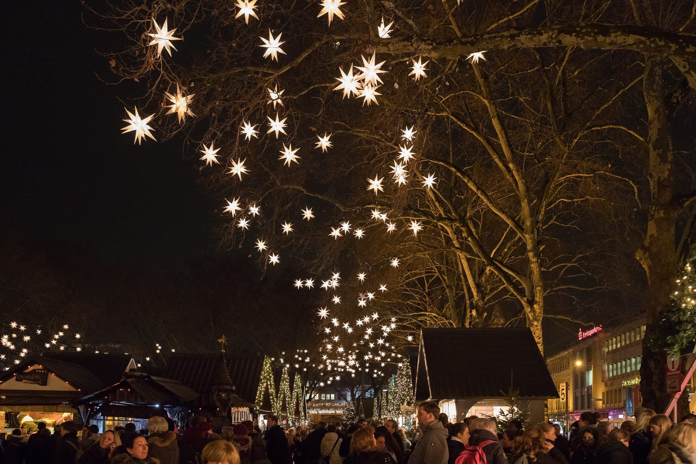 Crowds walk between the huts of the Markt der Engel under star lights in the Neumarkt trees