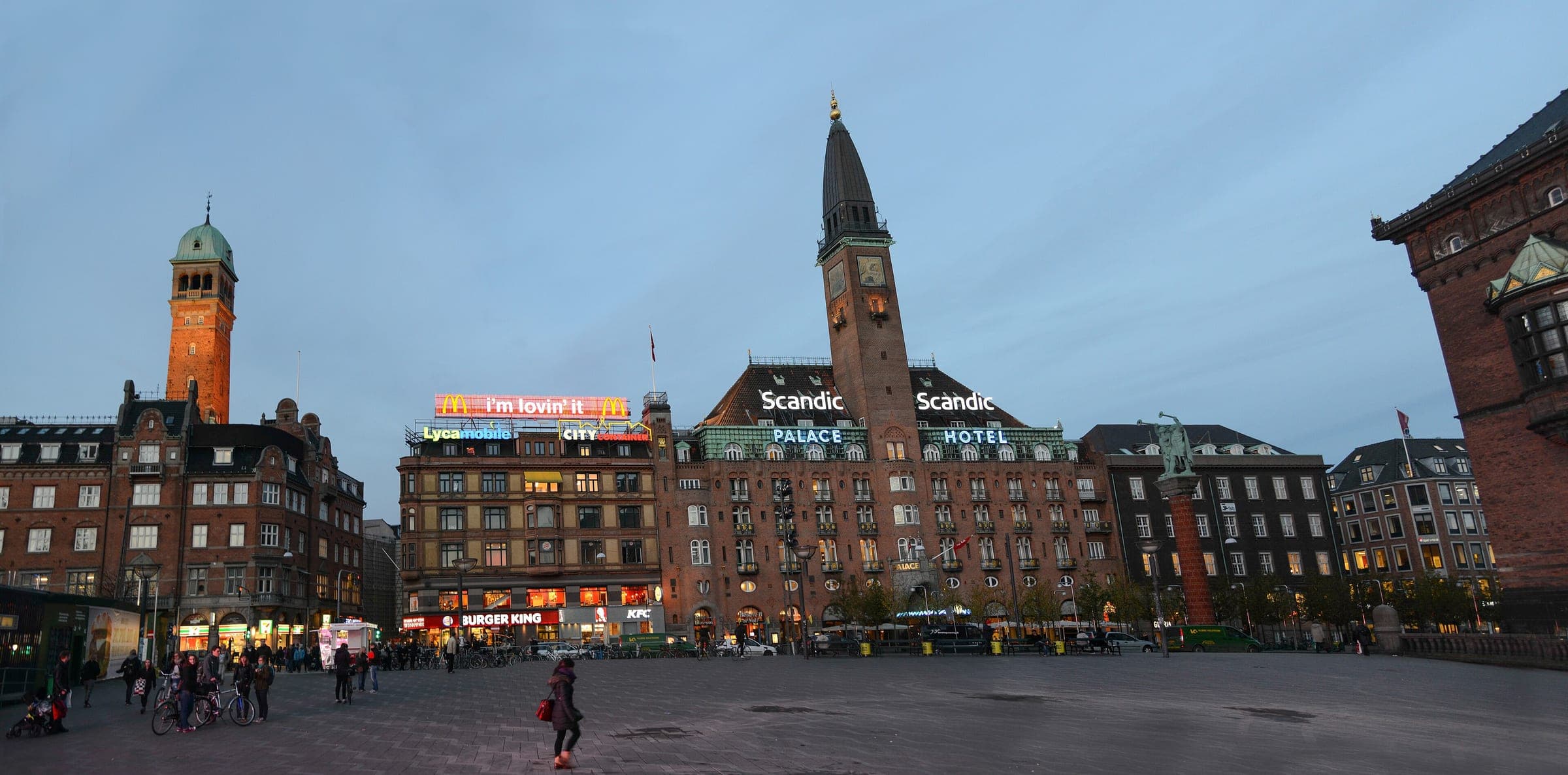 Shop signs light up around City Hall Square in Copenhagen at dusk below the tower of the Palace Hotel.