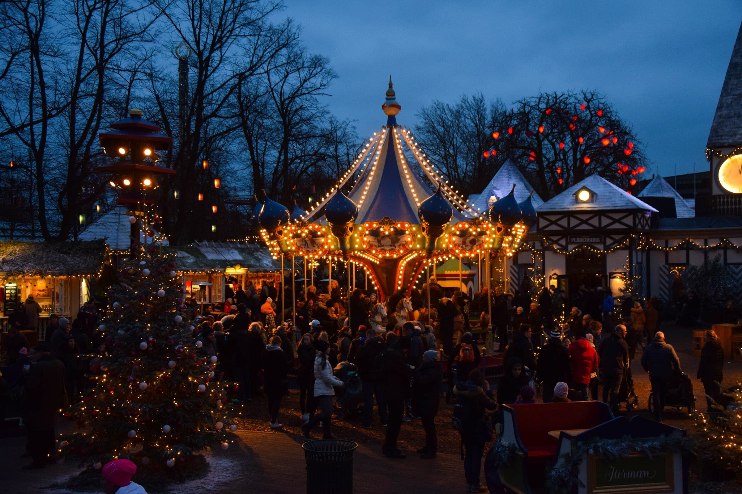 Visitors gather around a lit carousel among the Christmas stalls in Tivoli Gardens in Copenhagen at dusk.