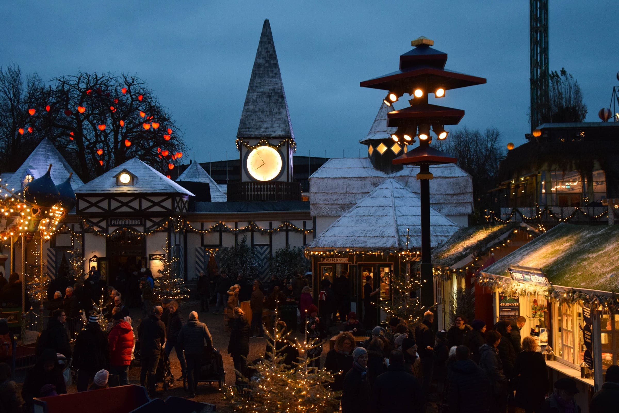 Crowds move between white-roofed stalls at Christmas in Tivoli in Copenhagen as blue hour settles over the garden.