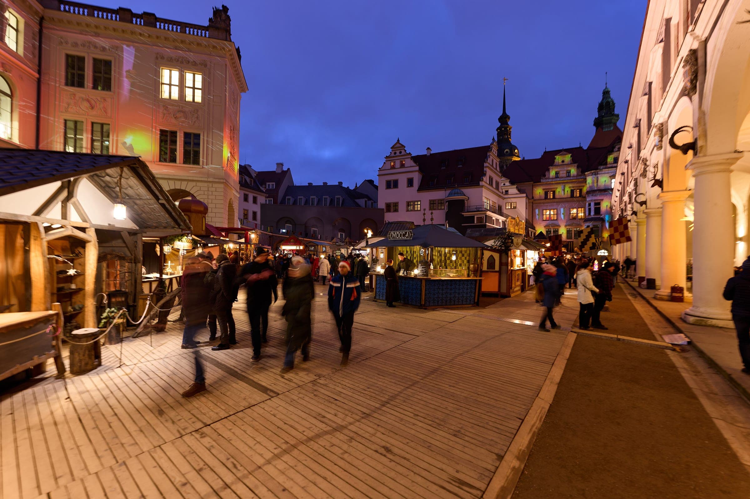 Visitors stroll past lit stalls in the Stallhof as dusk falls over the towers of Dresden's royal palace.