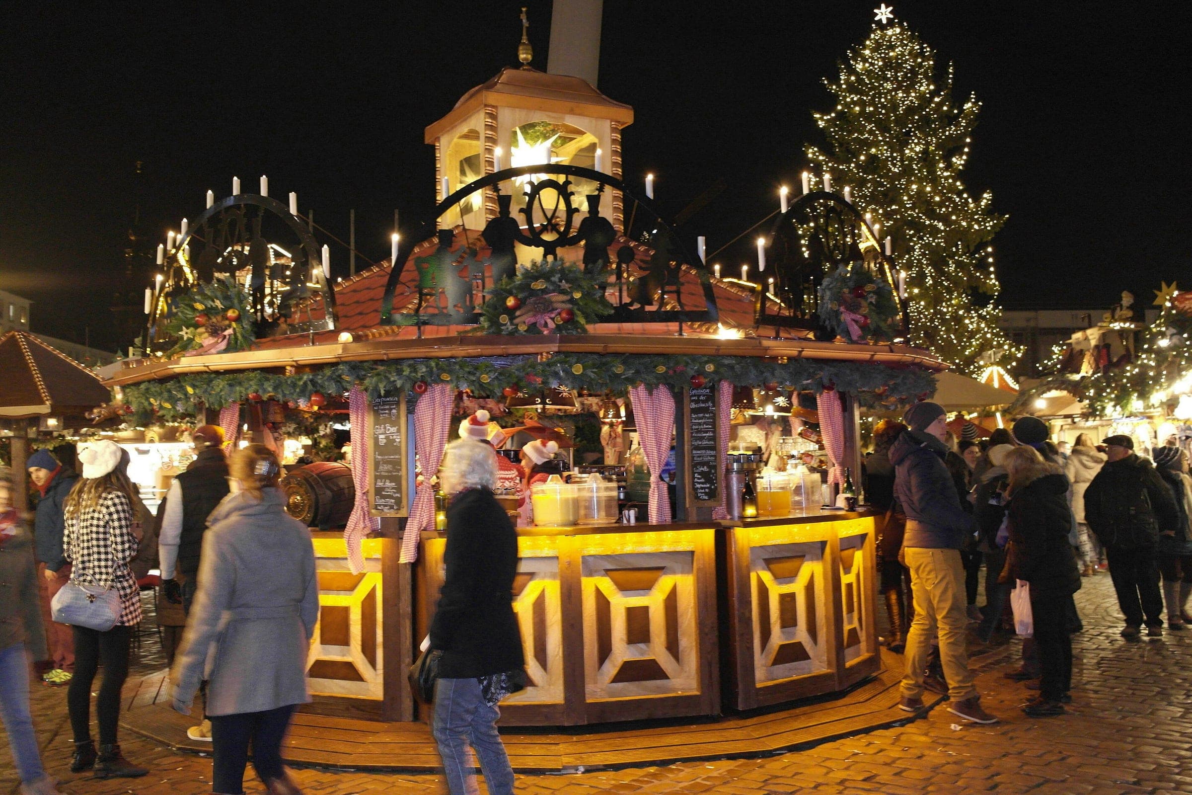 Visitors gather at a round mulled wine stall under a small pyramid at Dresden's Striezelmarkt at night.
