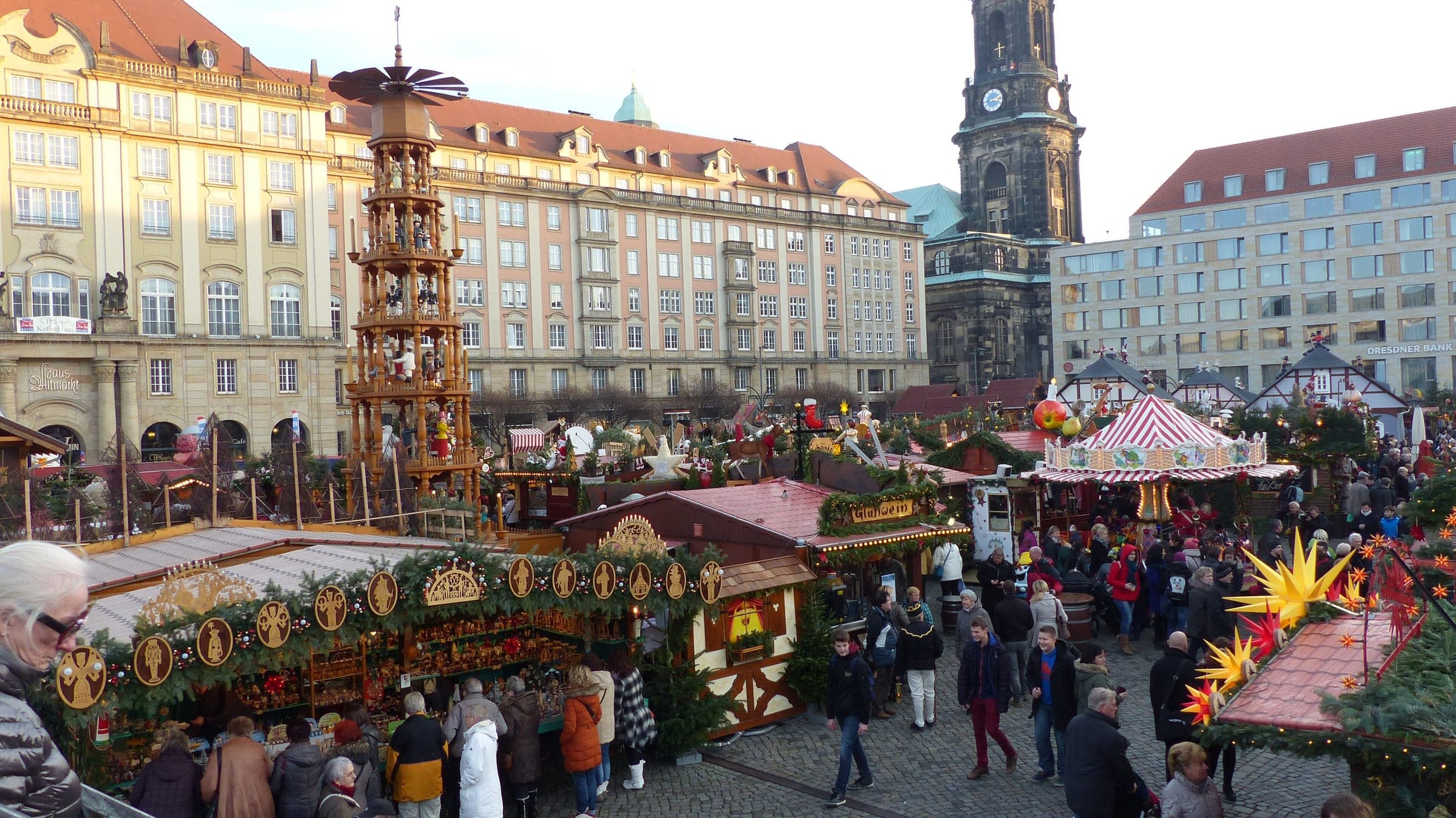 Crowds walk between the Striezelmarkt stalls on the Altmarkt in Dresden with the Kreuzkirche tower behind.