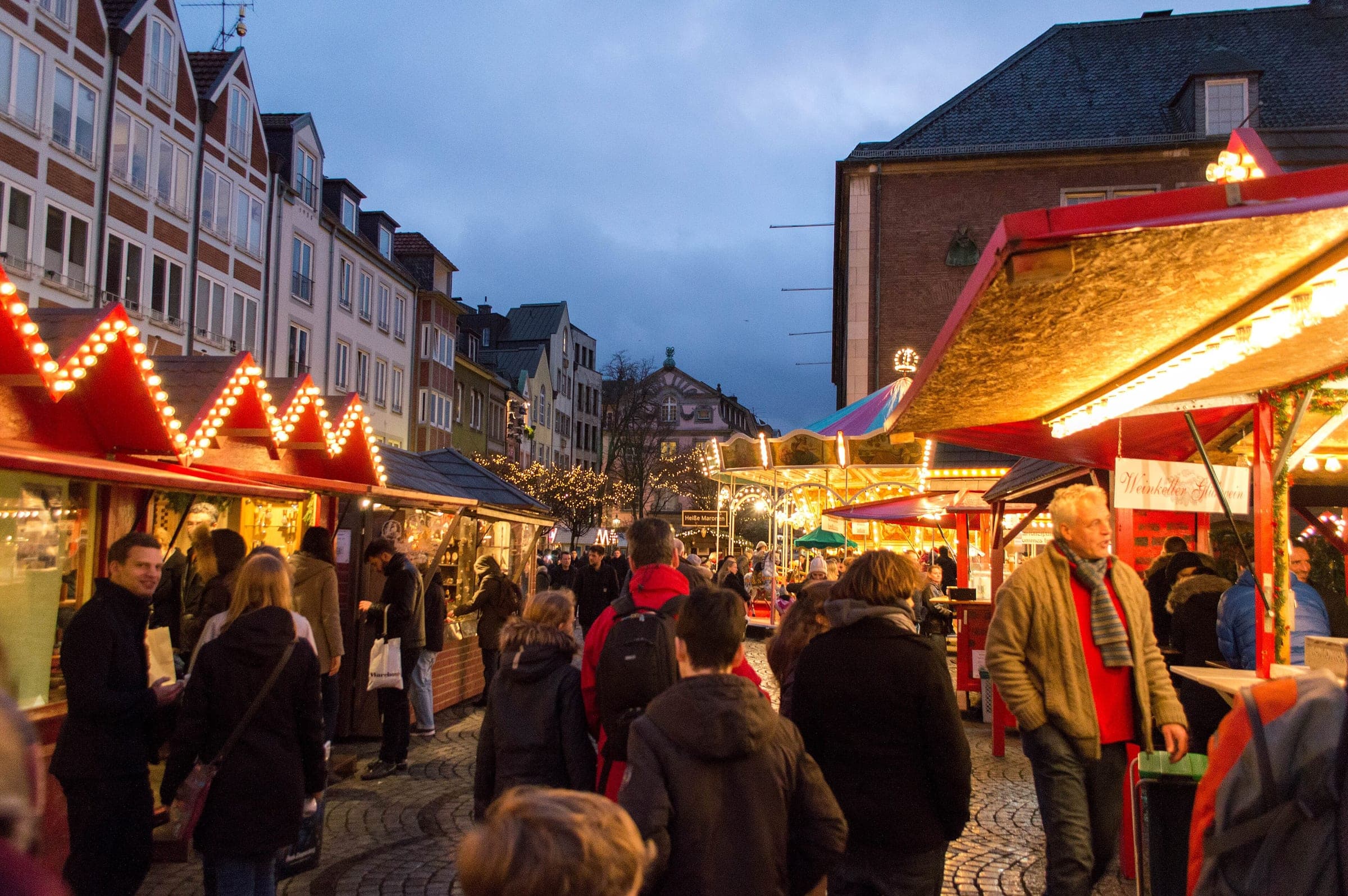 Shoppers stroll between lit stalls on Düsseldorf's Marktplatz in the Altstadt at dusk
