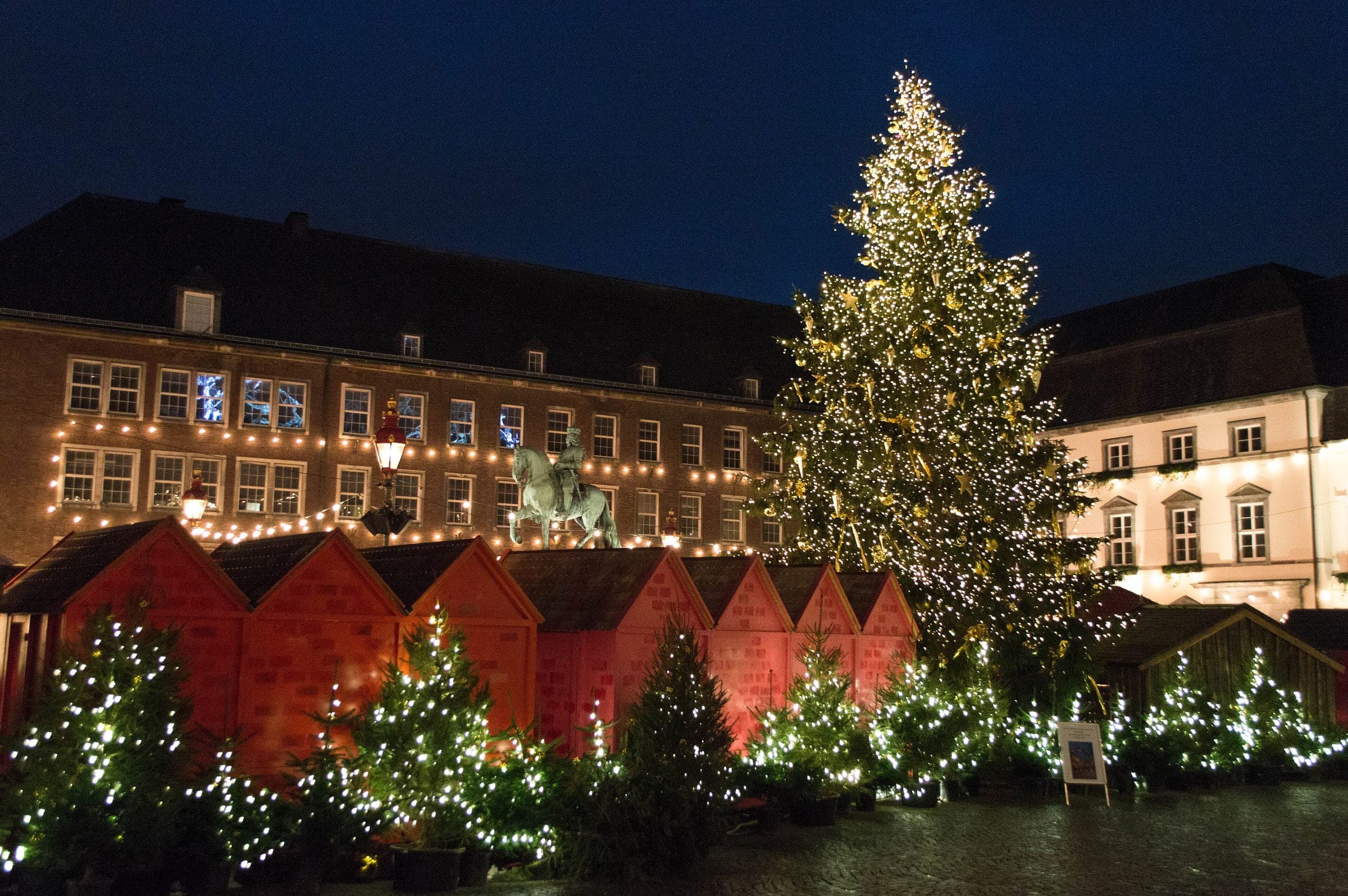 A tall lit tree and red huts fill the Marktplatz in front of Düsseldorf town hall at night