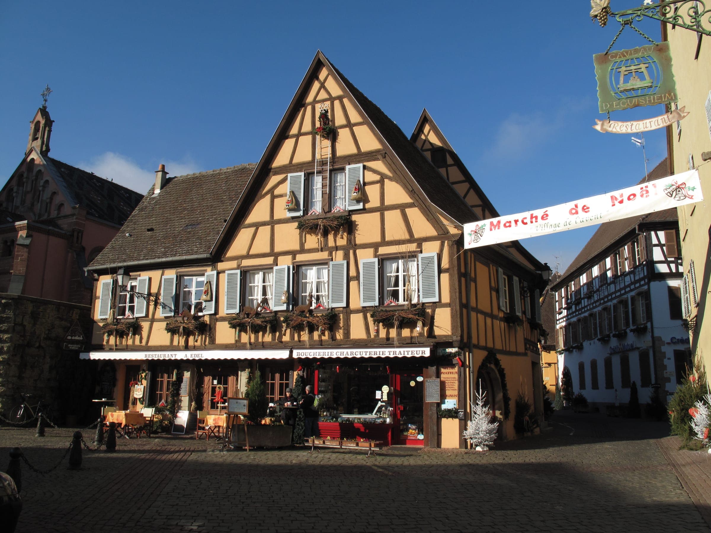 A Marché de Noël banner hangs beside a yellow half-timbered house on a square in Eguisheim.